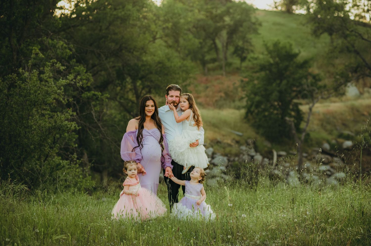 A family stands together in a lush green field with trees and rocks in the background during golden hour. Fresno Family Photography