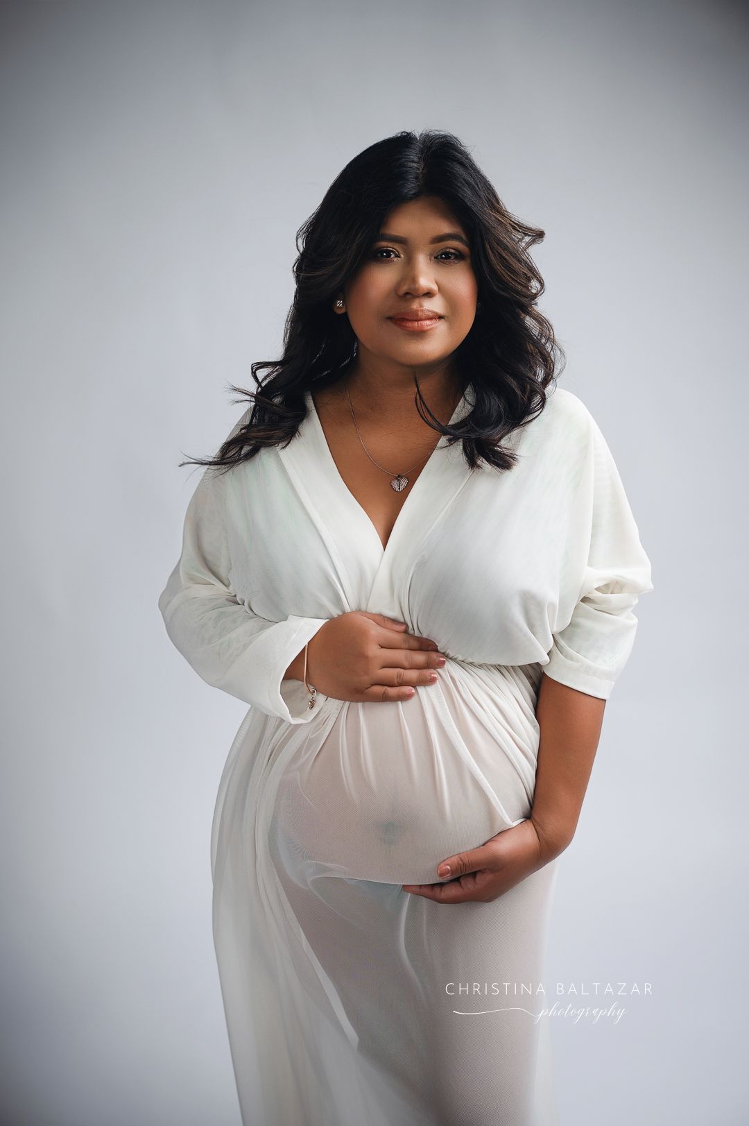 A pregnant woman in a flowing white dress poses against a gray background during a maternity photoshoot. Fresno Maternity Photographer