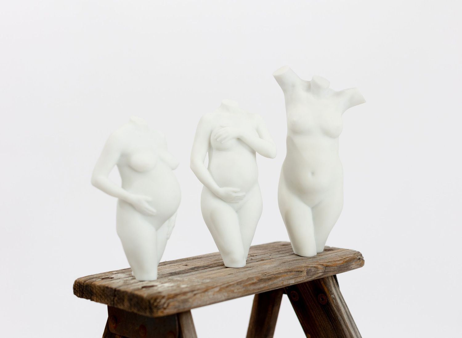 Three white ceramic pregnant female torso sculptures displayed on rustic wooden stool against white background. 3D Pregnancy Sculpture