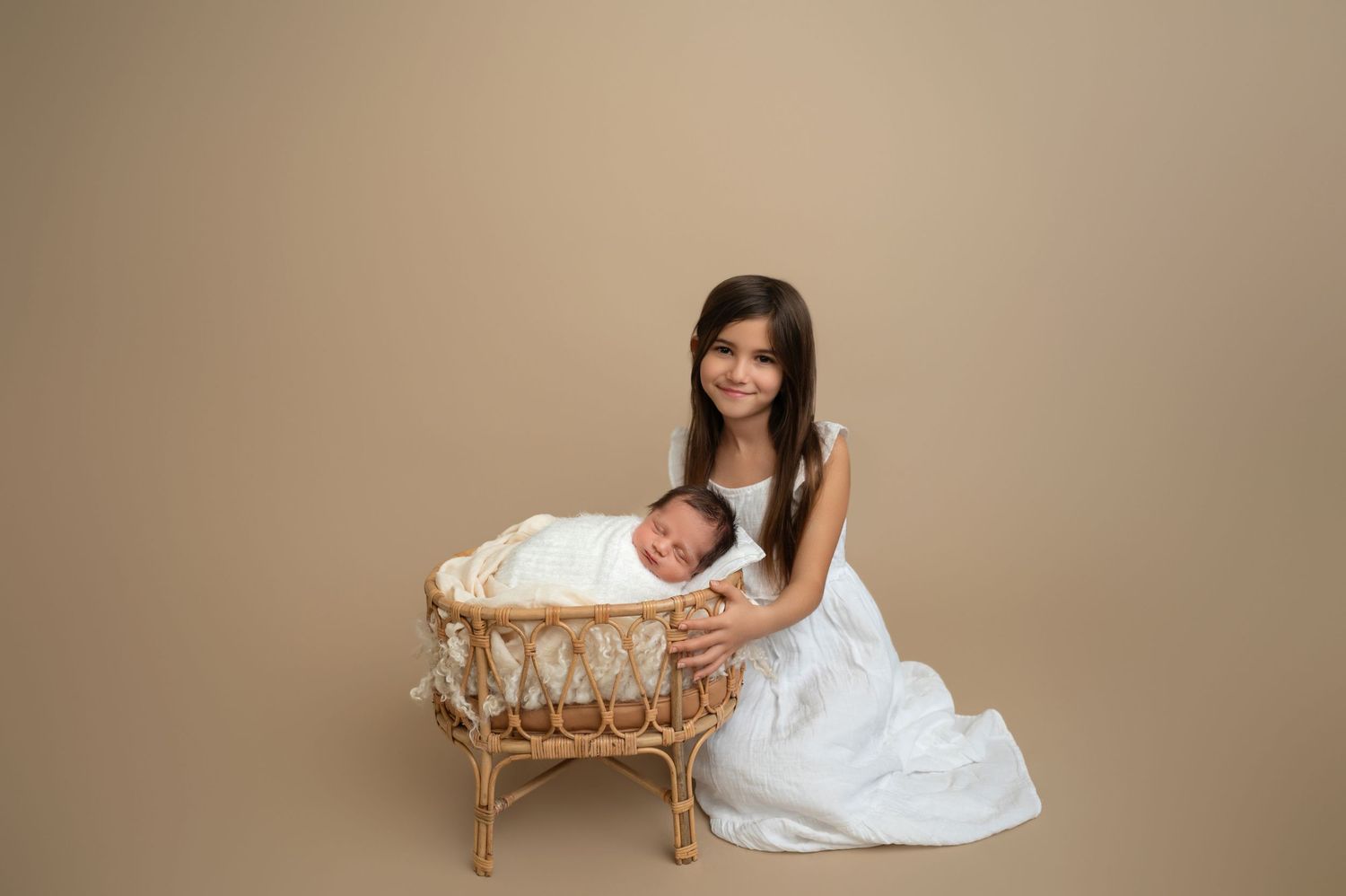 Portrait of child in white dress seated beside wicker bassinet with sleeping newborn.