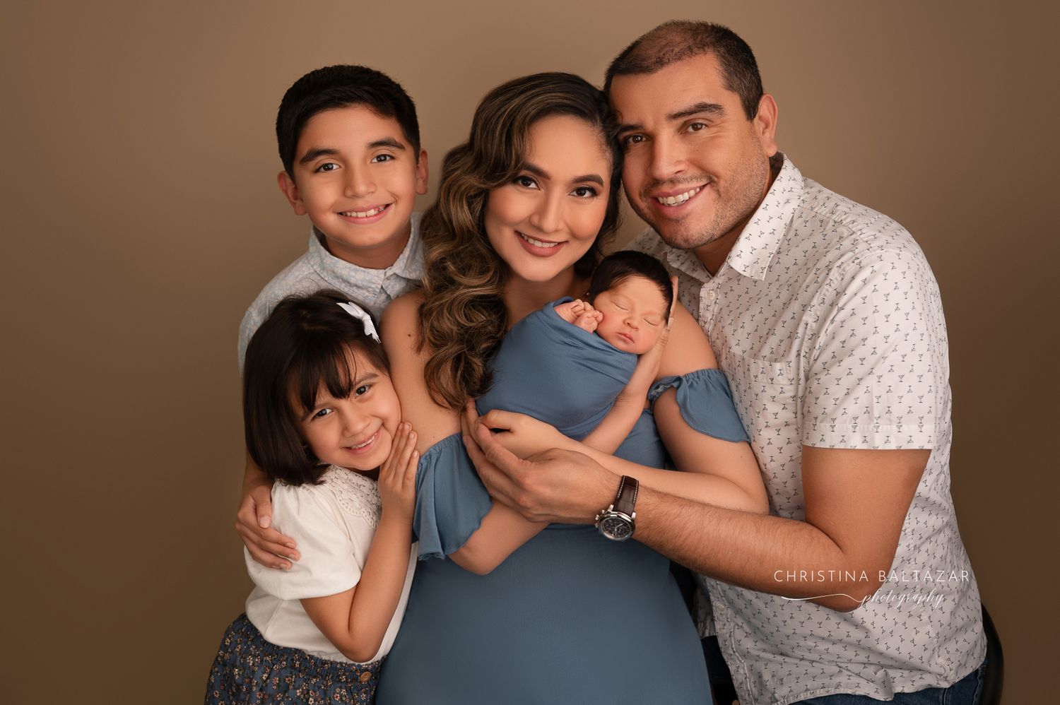A loving familyposes together against a brown backdrop for a warm studio portrait in coordinated casual attire. Fresno Newborn Photographer