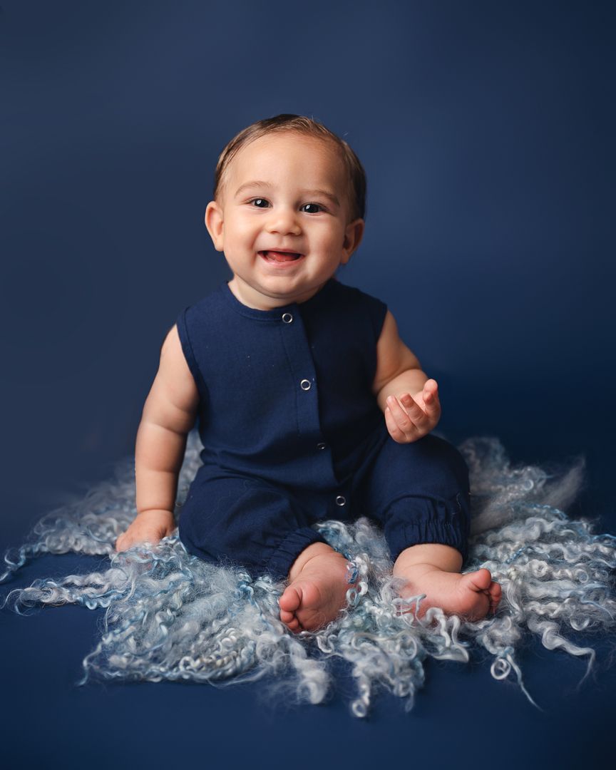 Baby in navy blue outfit posed on textured white fabric against dark blue backdrop.