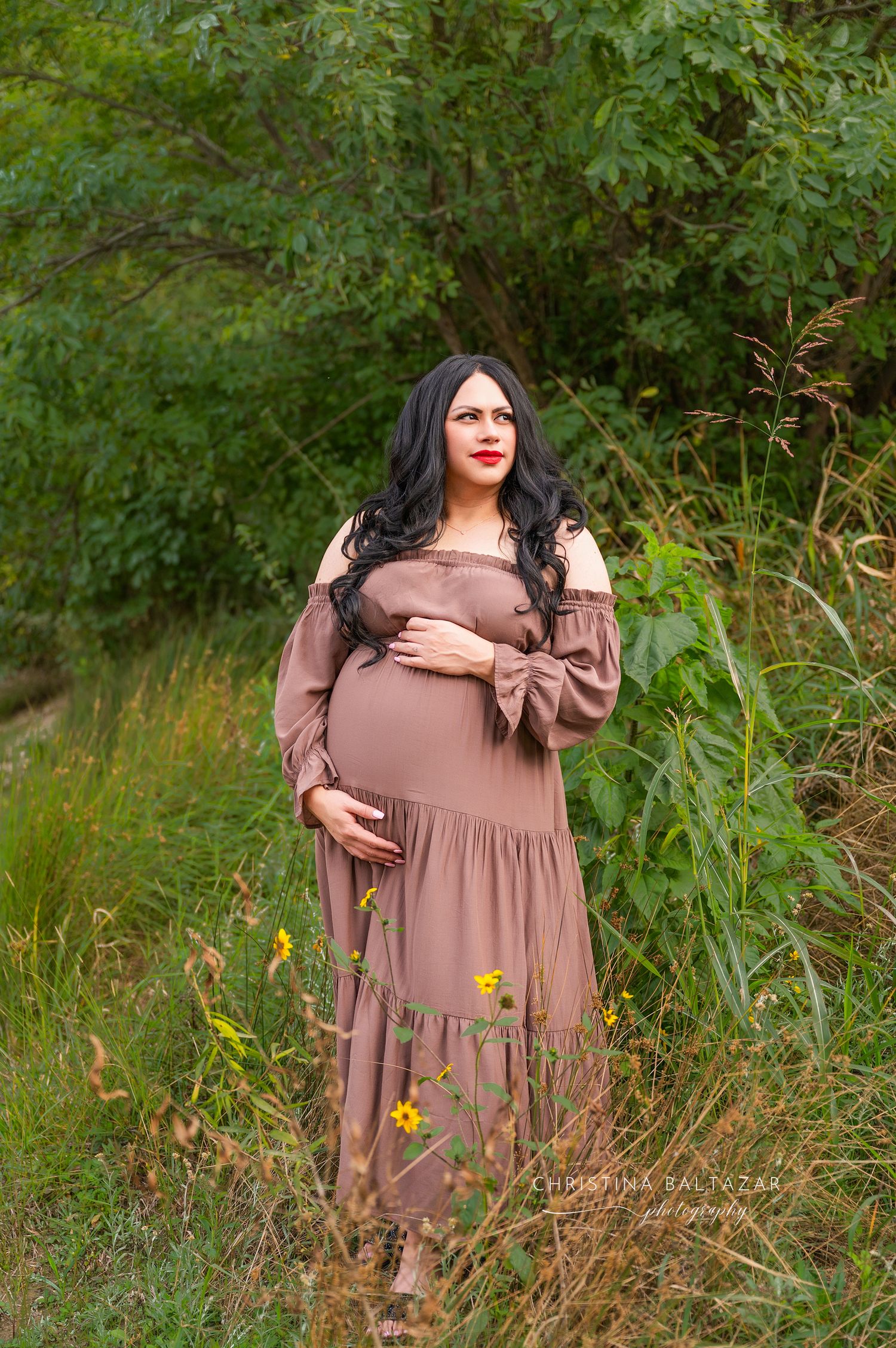 Expectant woman in flowing mauve dress poses outdoors among wildflowers and greenery in natural setting. Fresno Newborn Photography