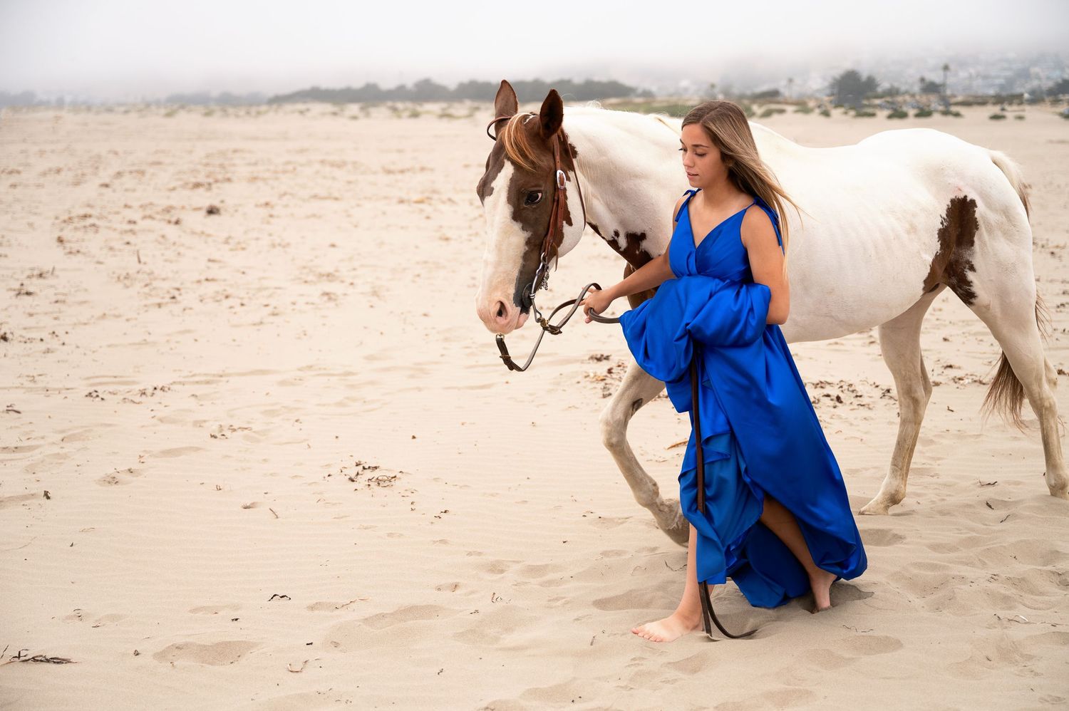 Blue formal dress contrasts with paint horse on misty beach setting.