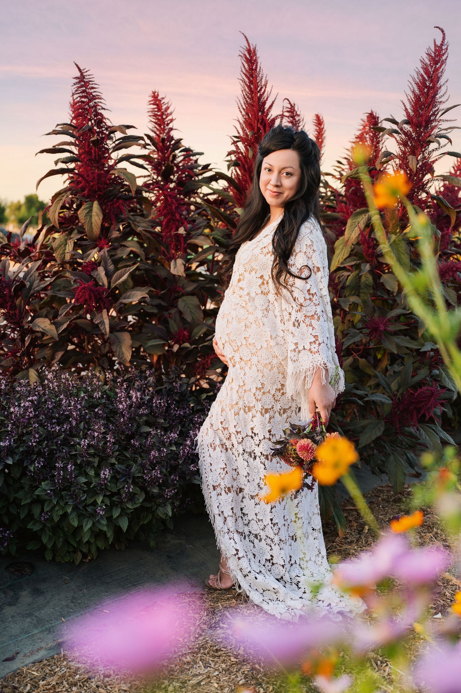 A white lace dress contrasts beautifully against colorful flowers and burgundy plants in a garden setting.