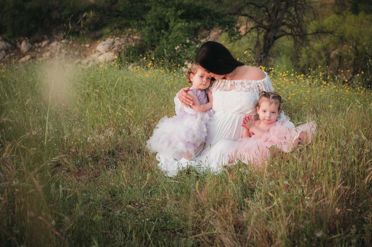Three figures in white and pink dresses sit together in tall summer grass. Fresno Family Photography