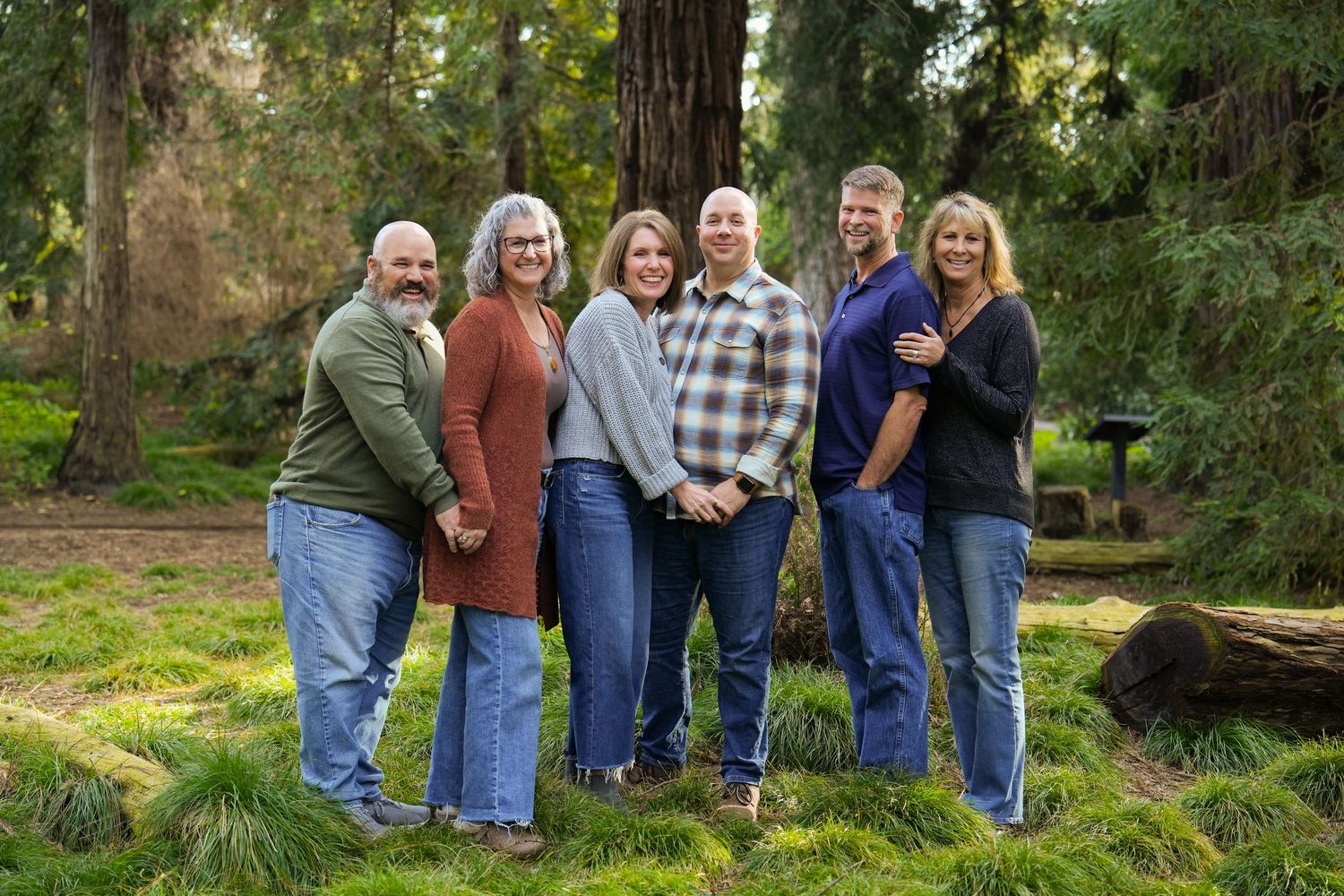 Six adults smiling together outdoors in a lush green forest park setting for a family portrait photo.