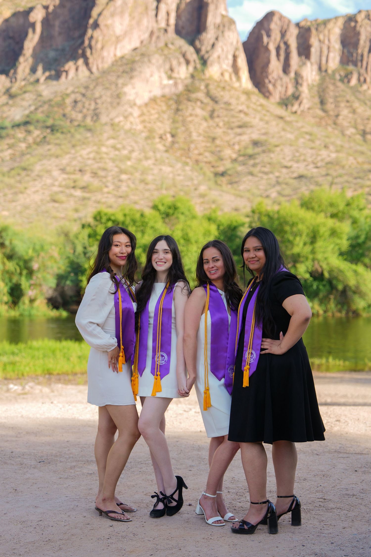 Four female graduates wearing purple sashes pose together outdoors near a river with scenic rocky mountains in the background.