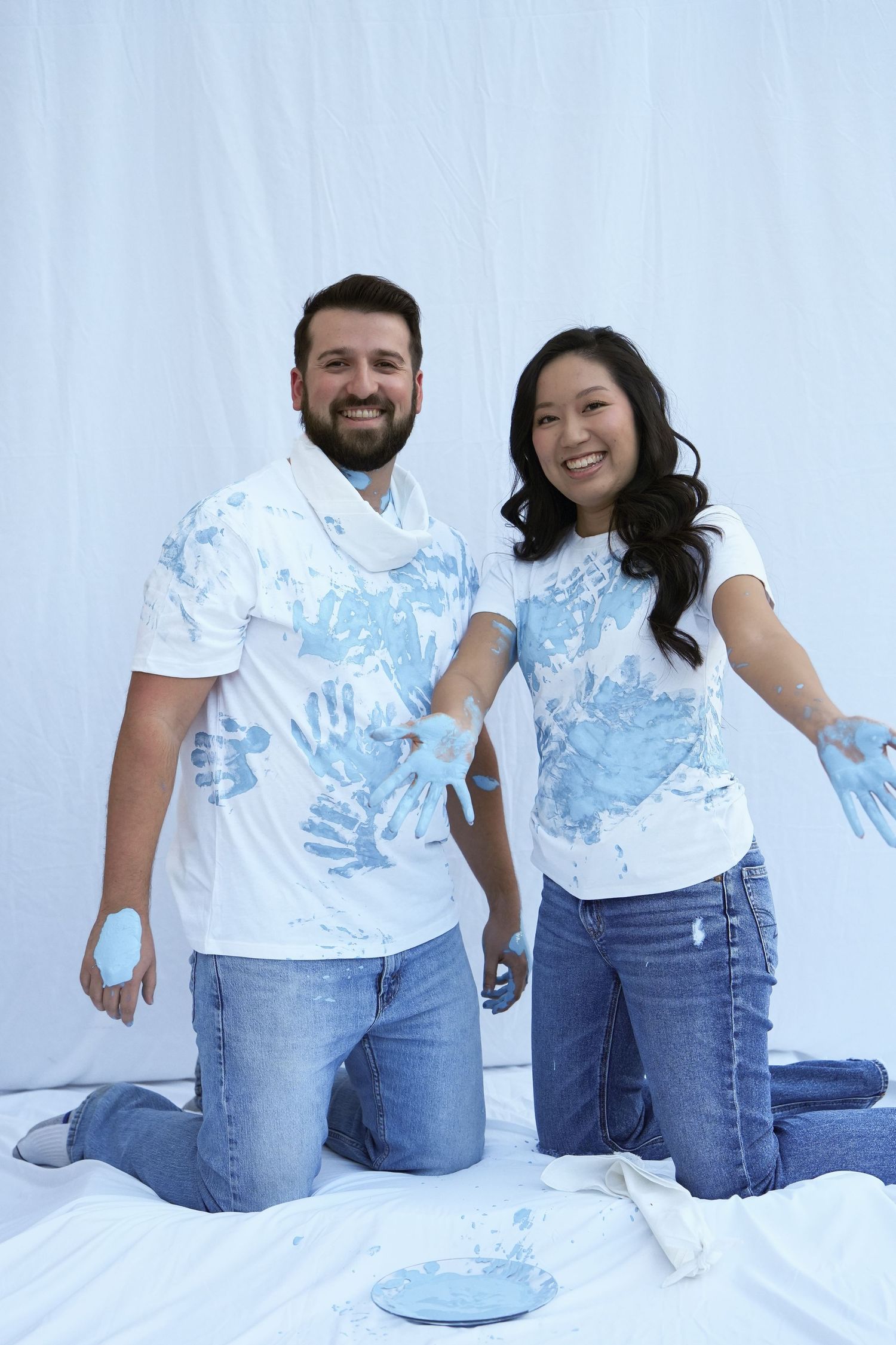 Two people wearing white t-shirts with blue paint handprints kneel on white fabric during a fun paint activity.