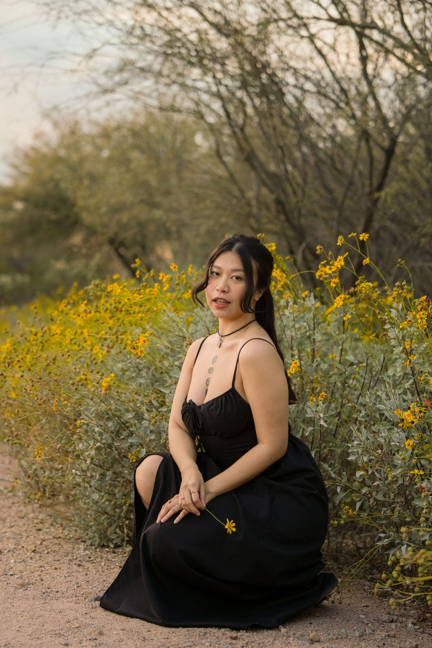 Woman in black dress sitting among yellow wildflowers in desert landscape, holding a small bloom.