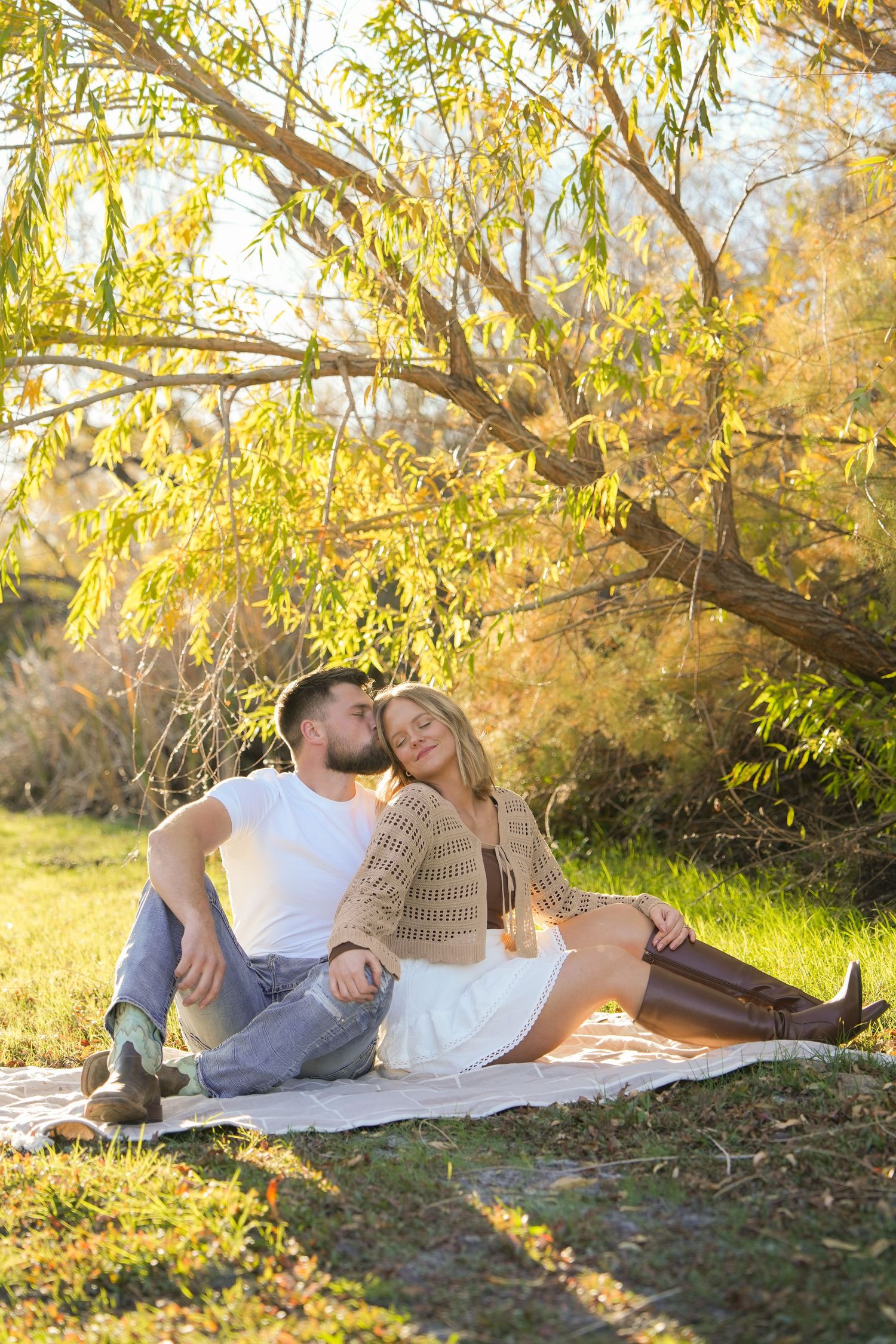 A couple relaxes together on a white blanket under golden willow trees during sunset in a park.