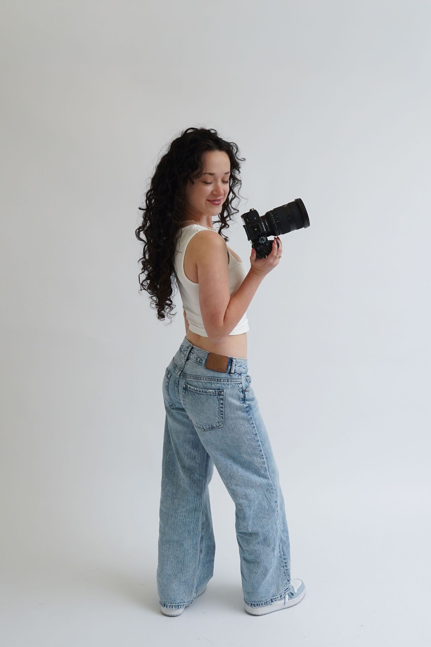 Young woman with curly hair holding a camera, wearing a white crop top and wide-leg jeans on white background.