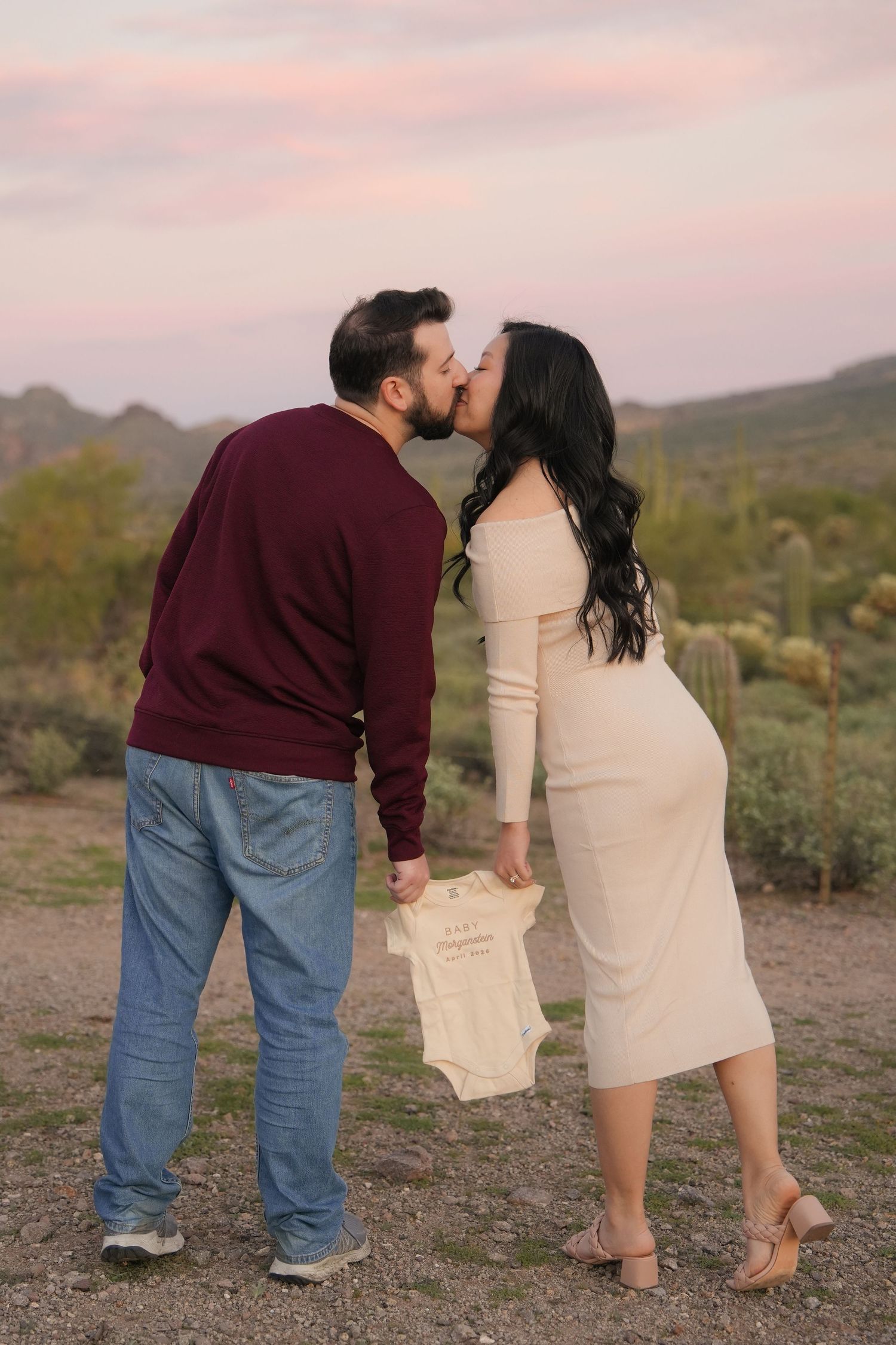 A couple shares a kiss at sunset in a desert landscape while holding a small baby onesie.