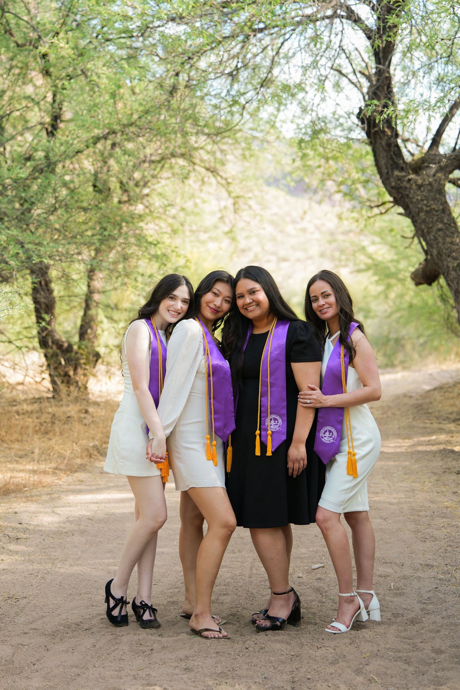 Four young women in graduation stoles pose together on a tree-lined dirt path celebrating their achievement.