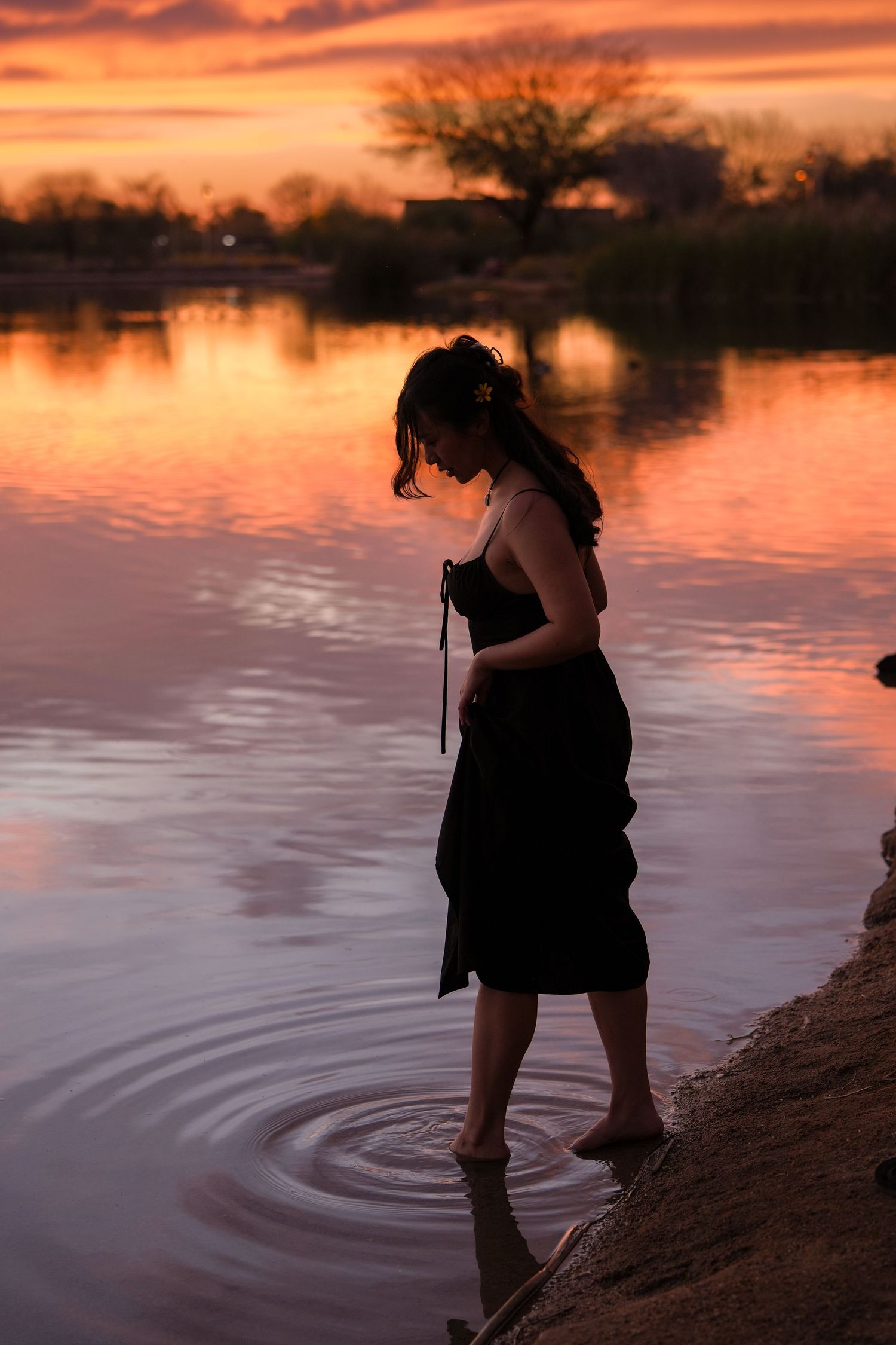 A silhouette stands at the water's edge during sunset, creating ripples in the calm lake as orange skies reflect on the surface.