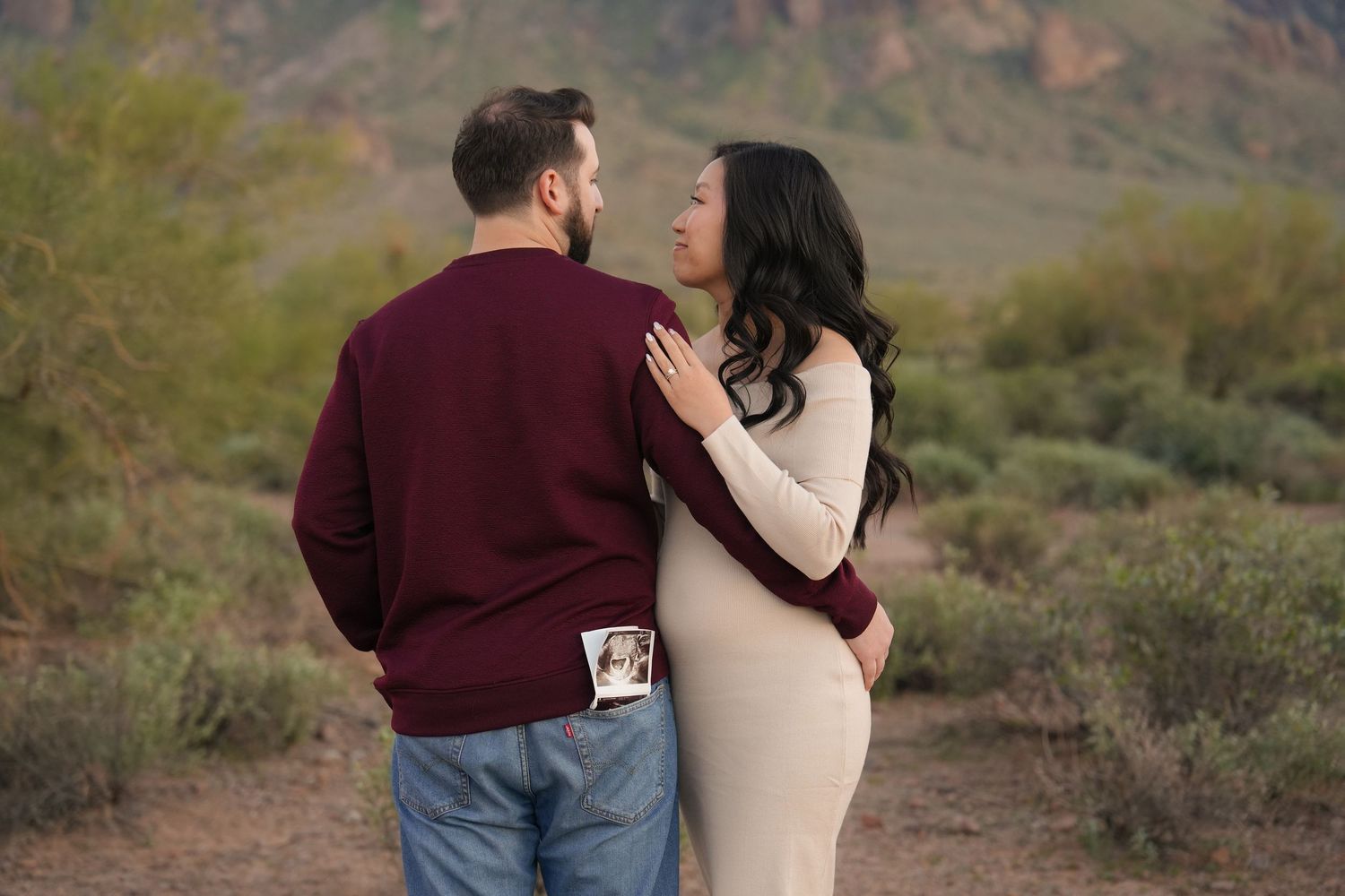 A couple in casual attire embraces on a desert trail with mountains in the background during golden hour.