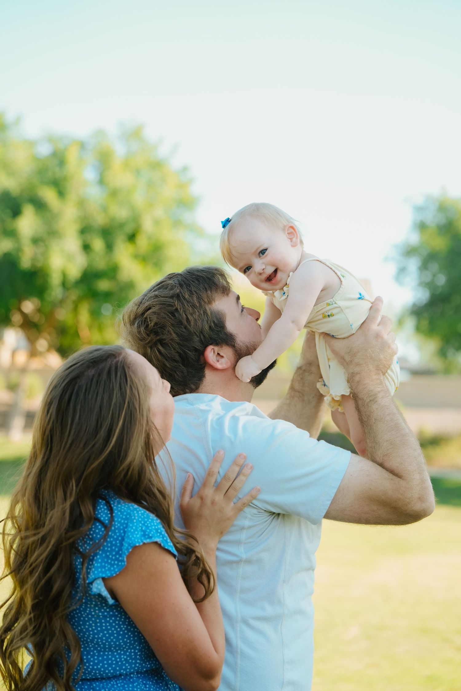 Happy family outdoors, parents lifting smiling baby girl in sunny park setting.