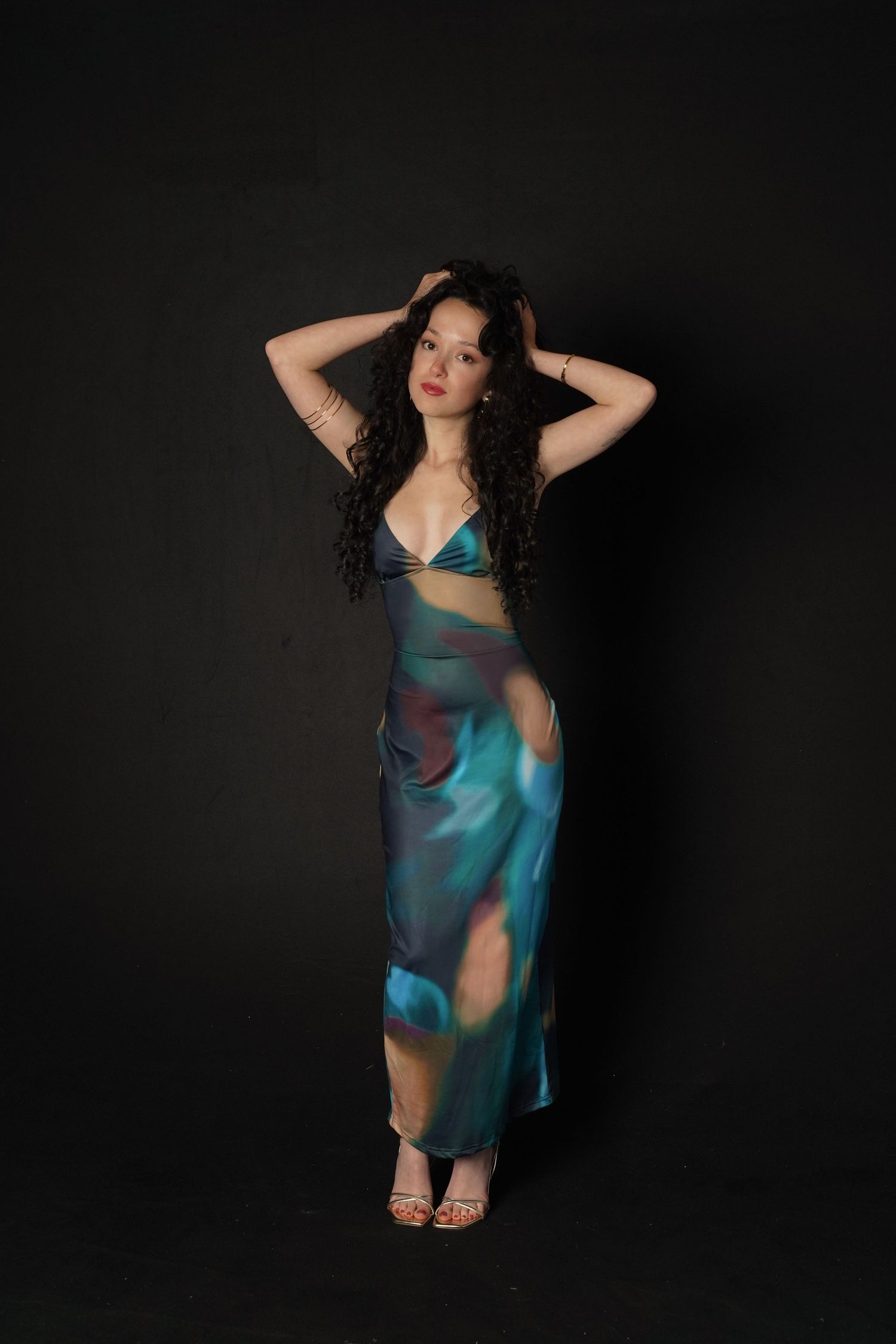 Woman in colorful teal satin slip dress with hands in hair, posing against dark studio background.