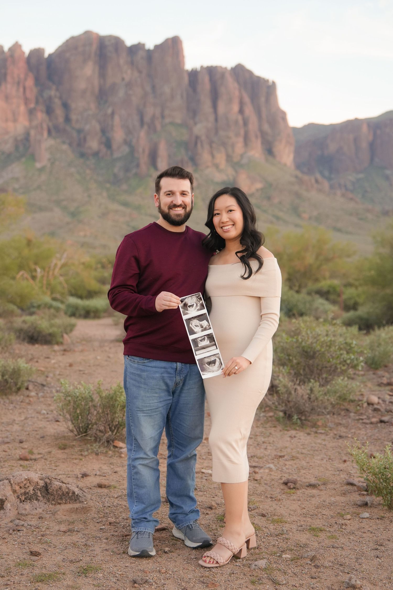 Expectant couple holding ultrasound photos in front of desert mountain landscape.