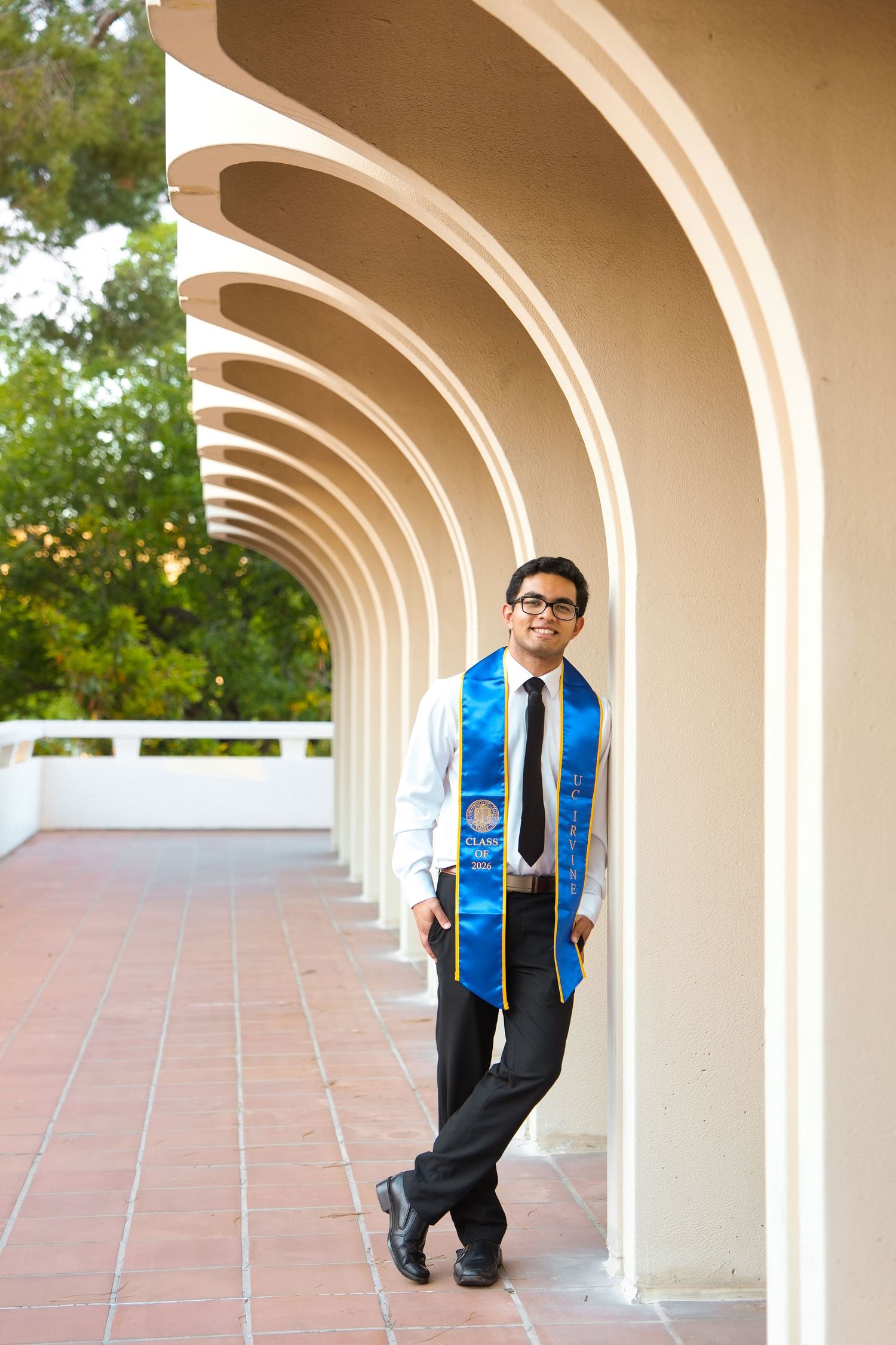Young man in graduation stole and formal attire posing confidently under arched colonnade walkway.