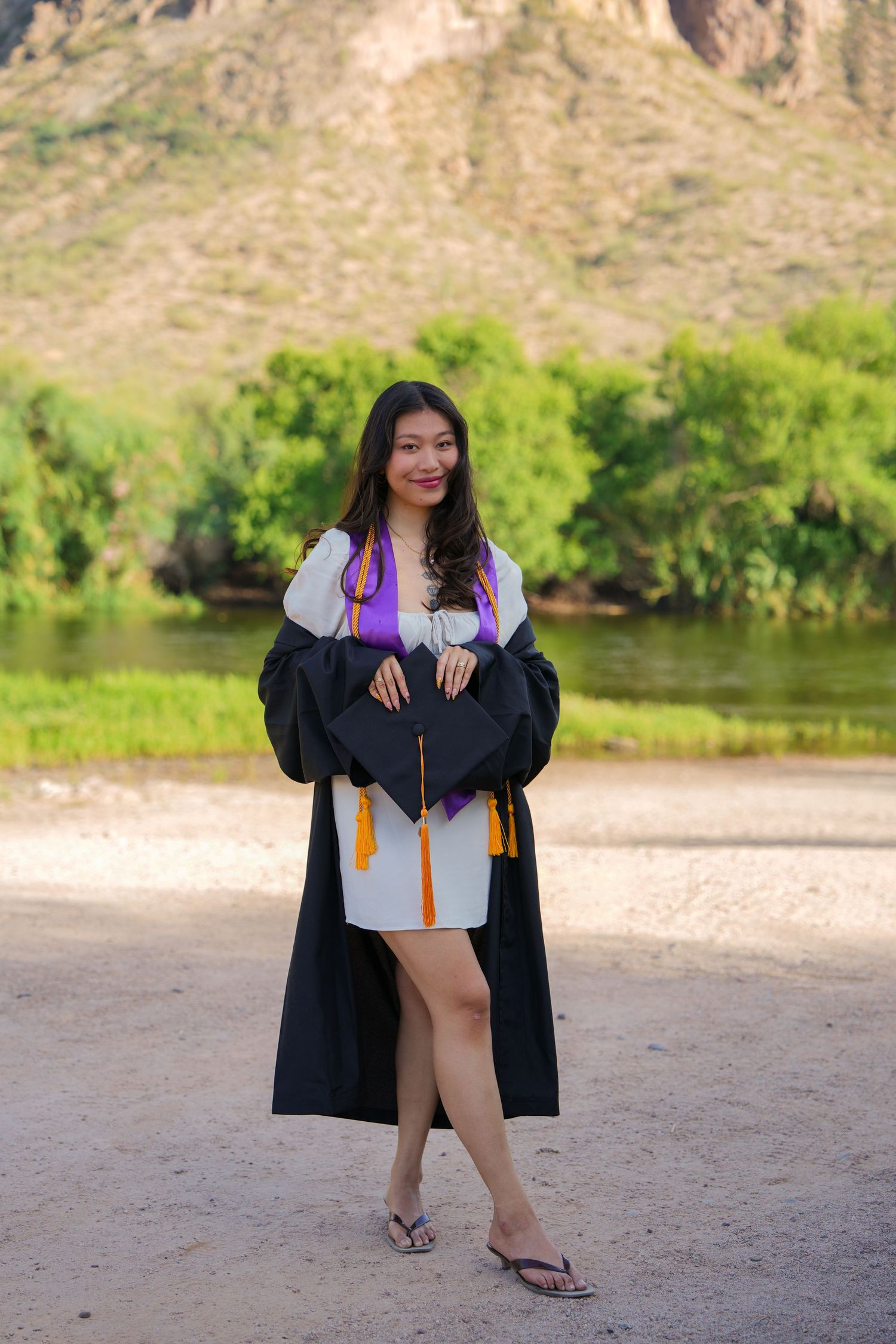 Young woman in graduation gown and cap with purple stole poses outdoors near river with mountain backdrop.