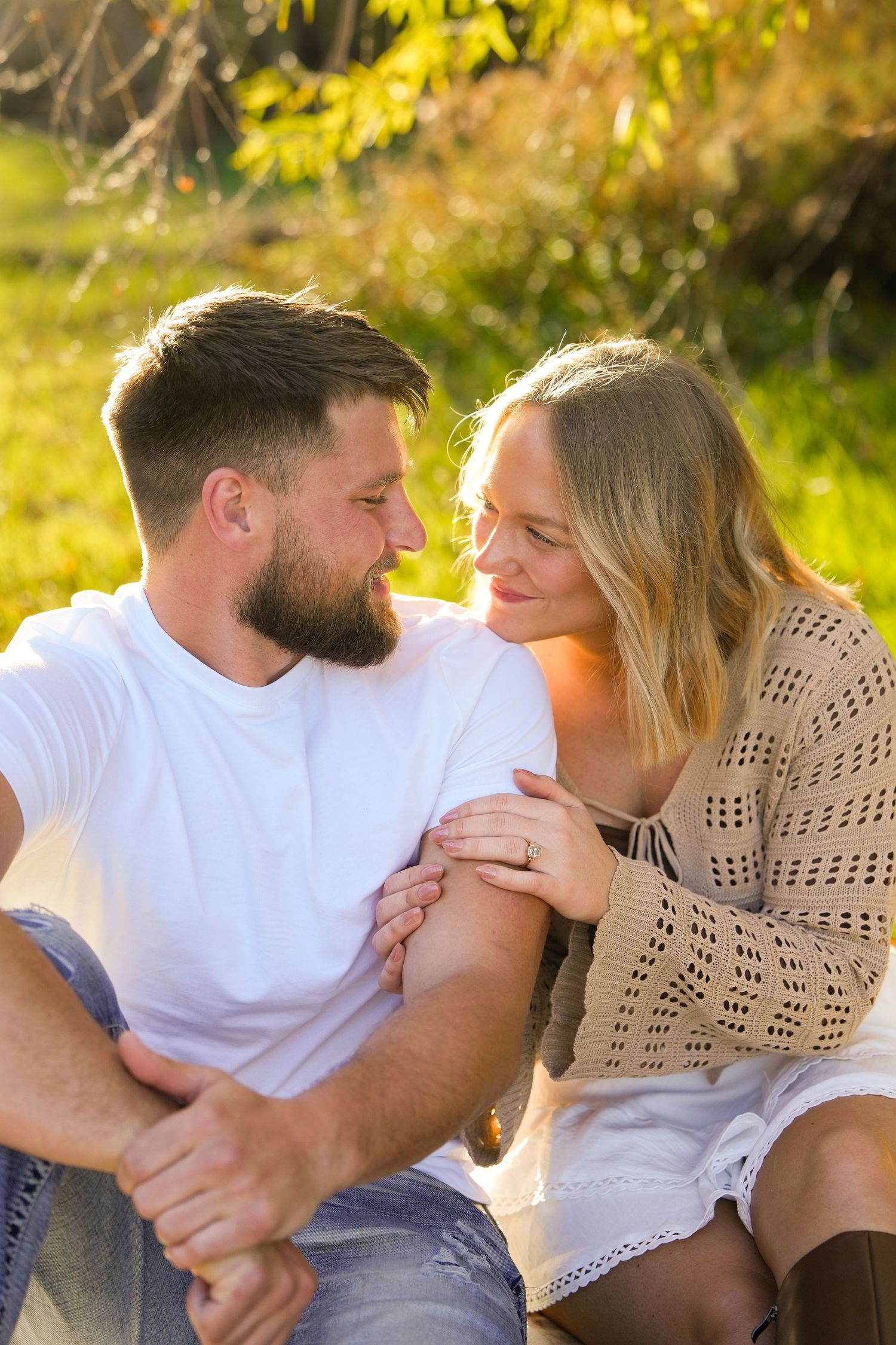 A couple sitting outdoors in golden sunlight, smiling at each other while holding hands in a lush green park setting.