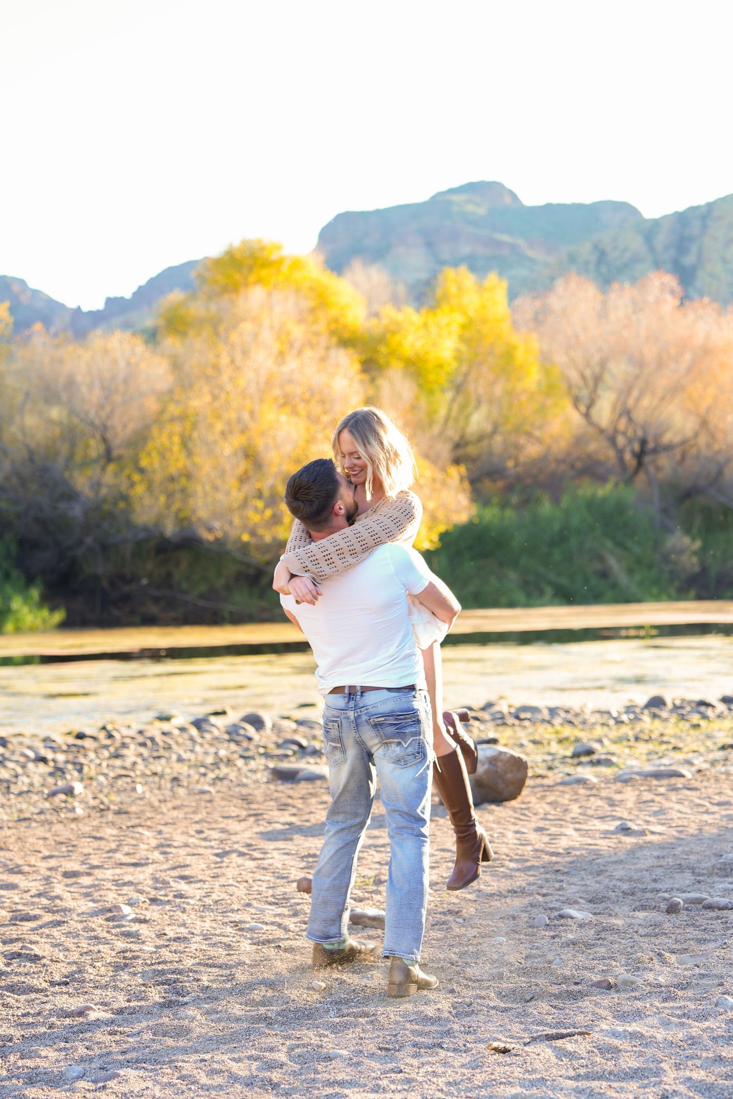 A couple embraces and spins playfully on a sandy beach with autumn foliage and mountains in the background.