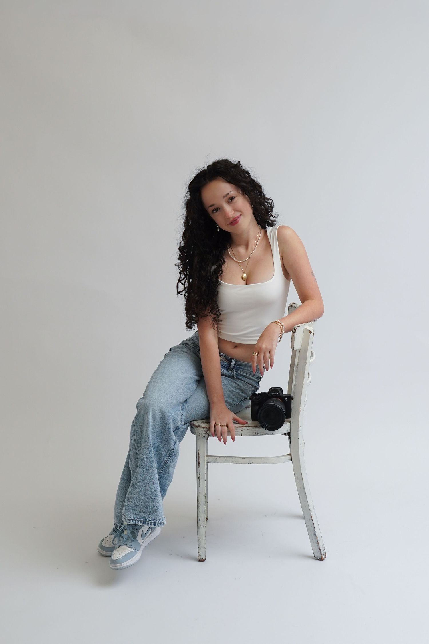 Young woman with curly hair sits on white chair wearing crop top and jeans, holding a camera in a studio setting.