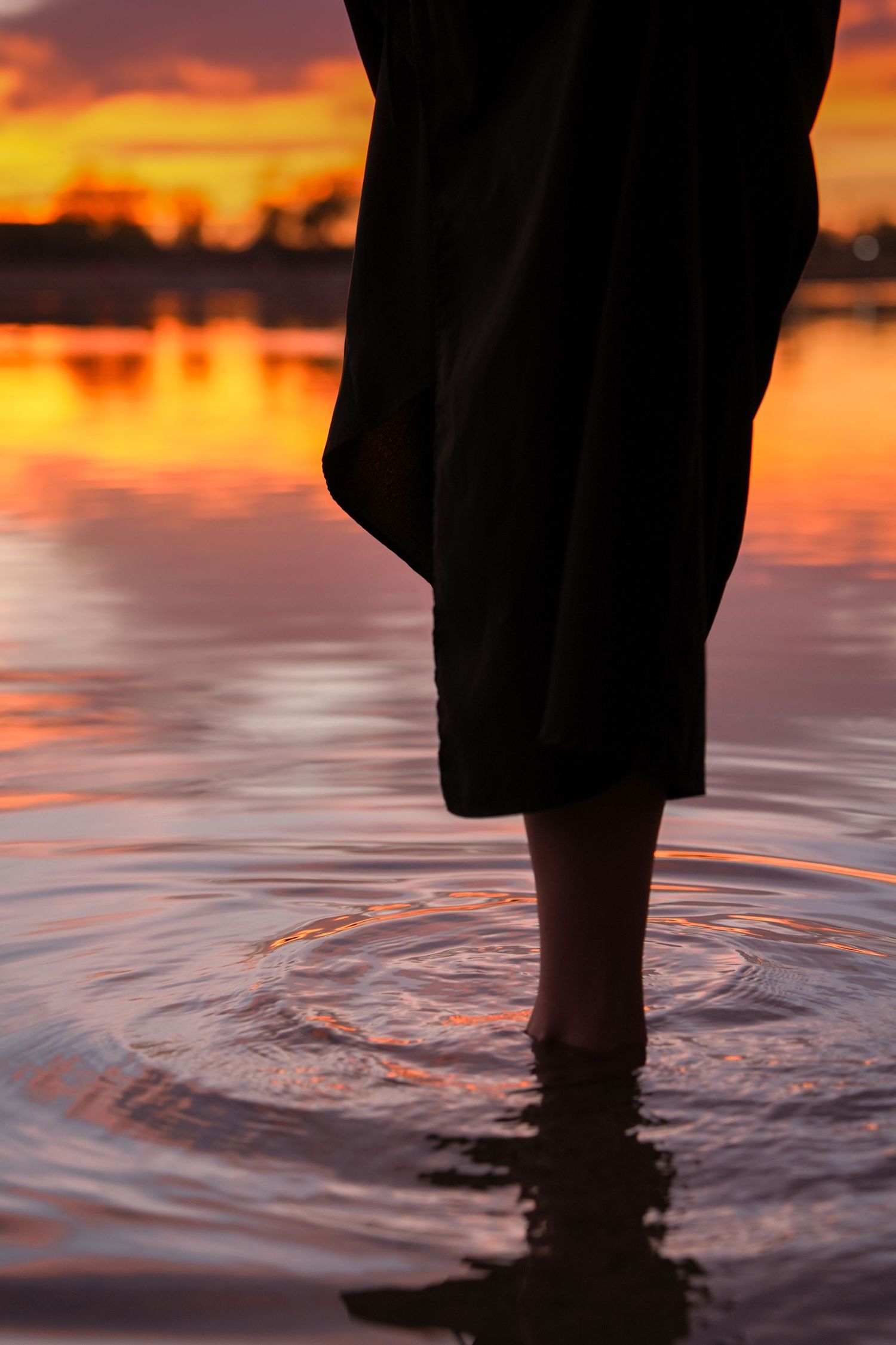 Person's feet standing in calm water at sunset with vibrant orange sky reflecting on the rippling surface.