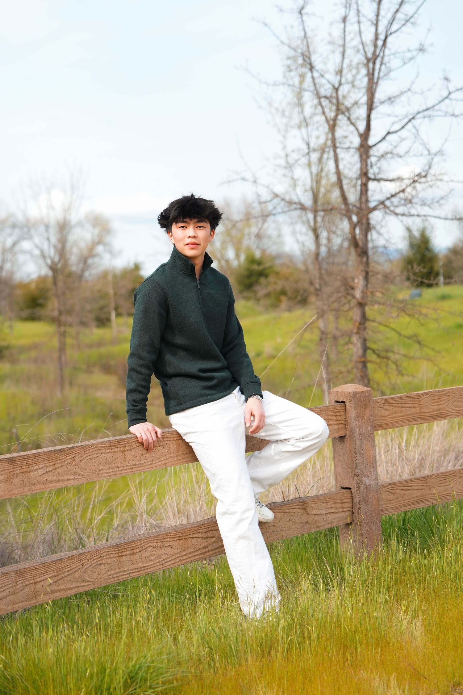 Person in dark sweater and white pants sitting on wooden fence in grassy field.