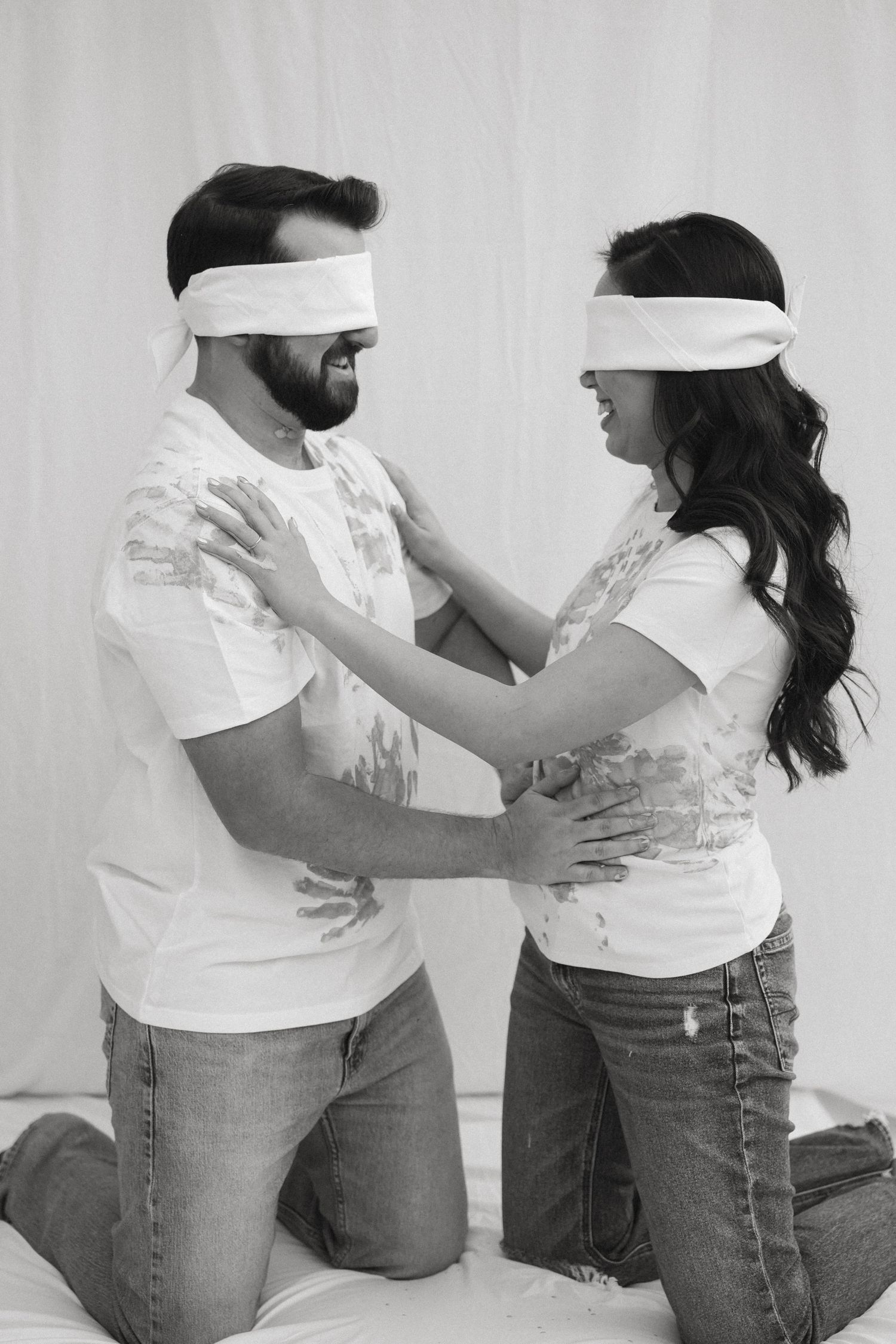 A couple wearing white blindfolds and shirts kneel facing each other in an intimate black and white portrait.
