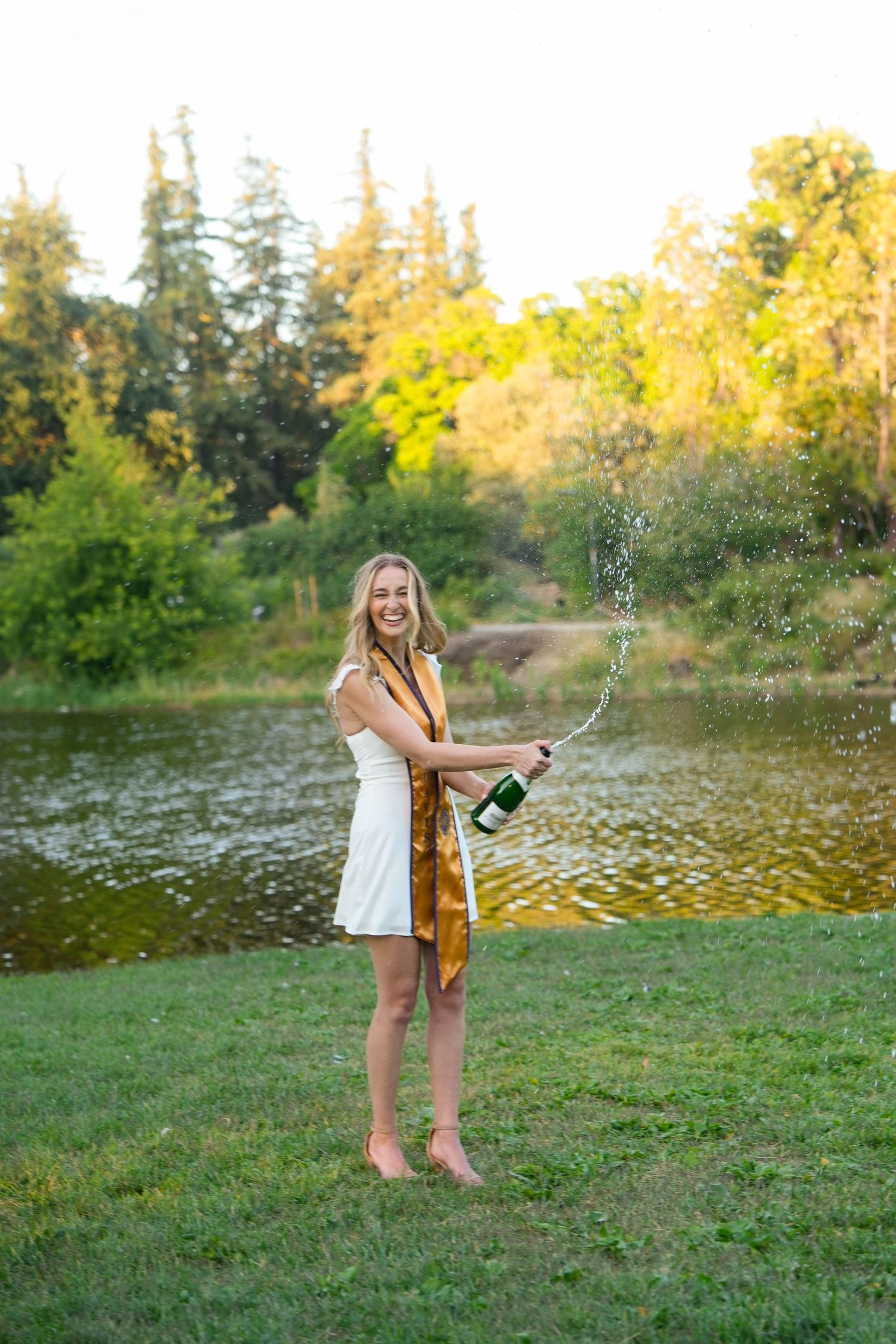 Young woman in white dress popping champagne bottle by a serene pond surrounded by lush green trees.