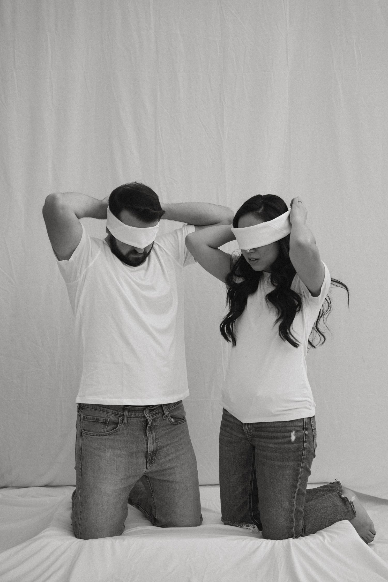 Two people in white t-shirts and jeans pose with their backs turned to the camera in a minimalist setting.