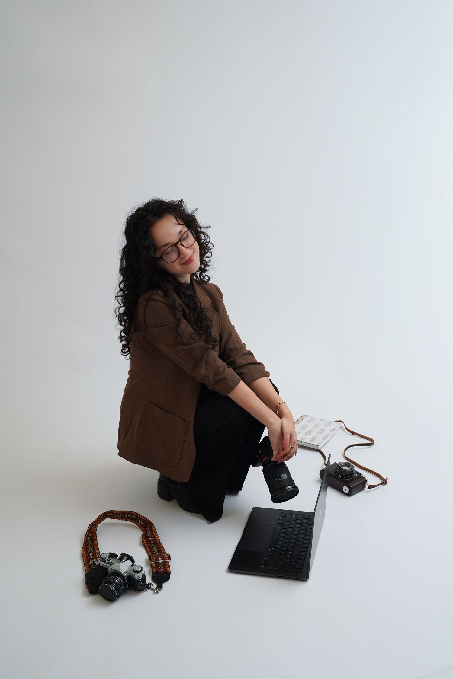 Woman photographer sitting on floor with DSLR camera, laptop, and film cameras on white background.