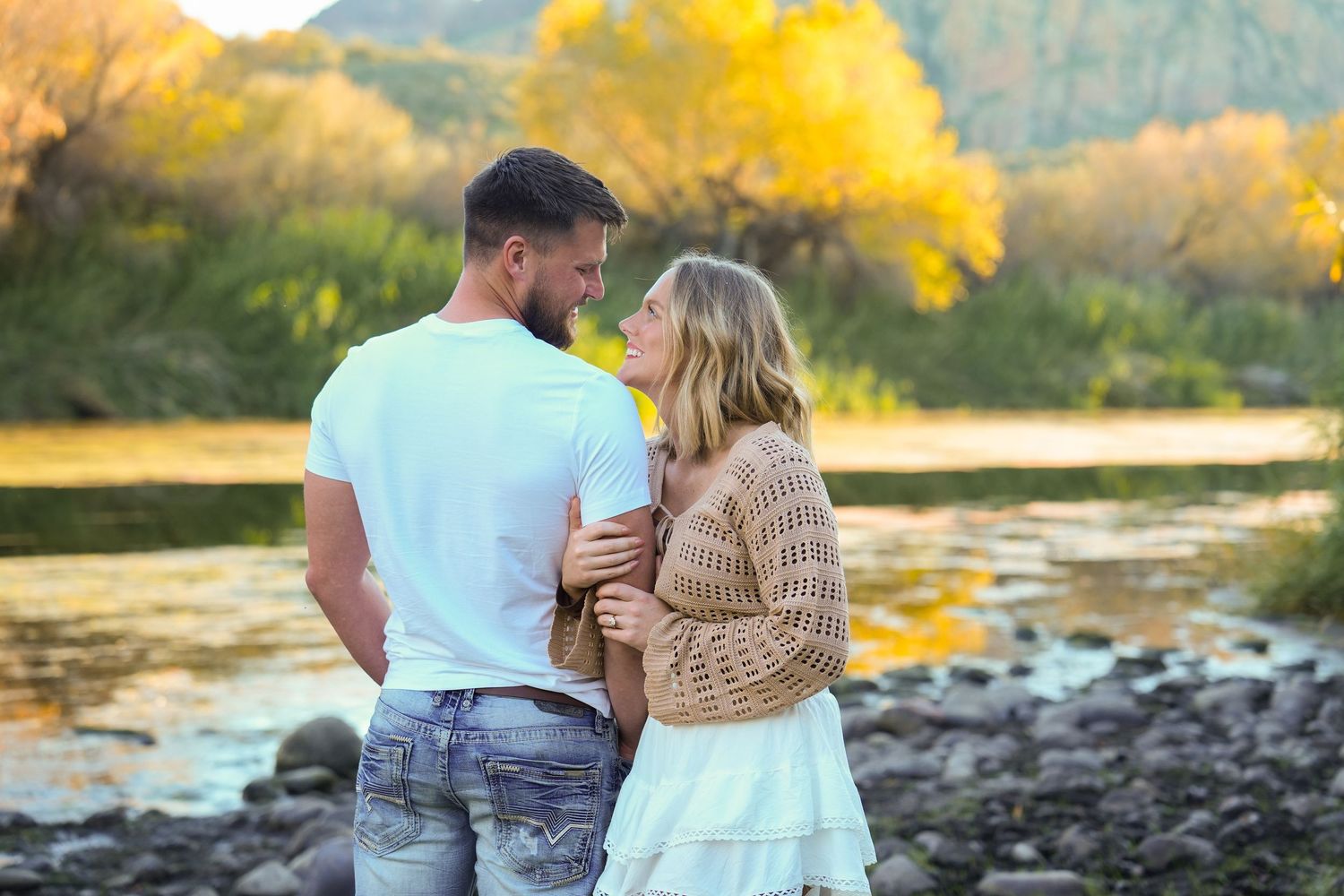 A couple shares a romantic moment at sunset by a river with golden autumn trees in the background.