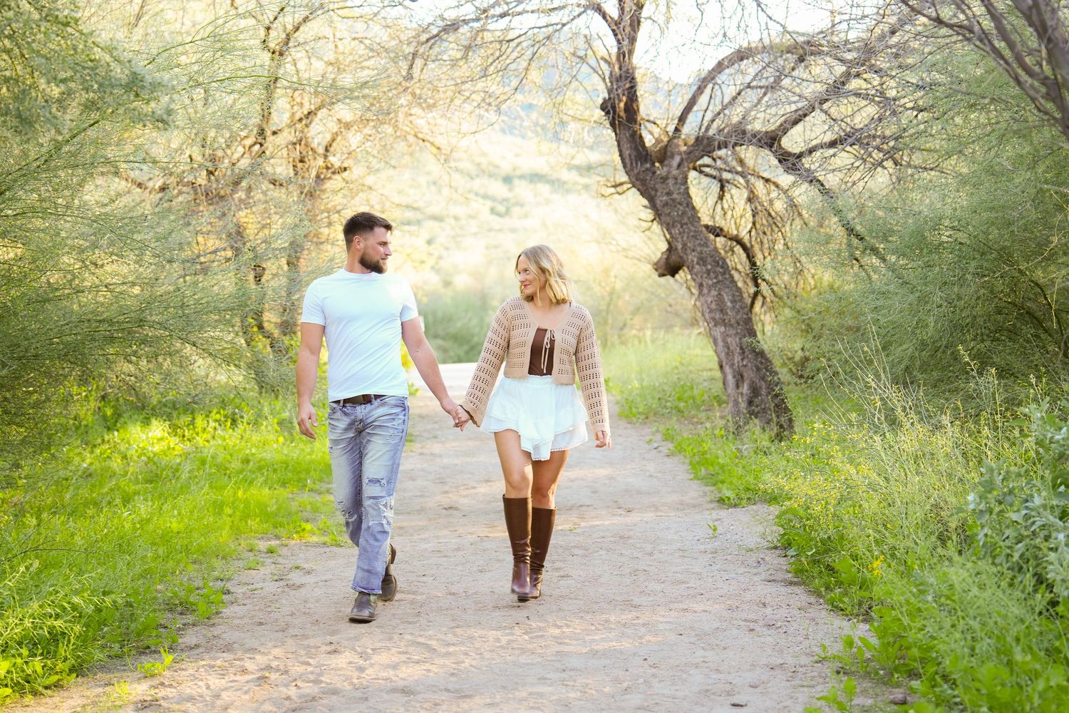 A couple walks hand in hand down a sunlit nature path surrounded by lush green foliage and trees.