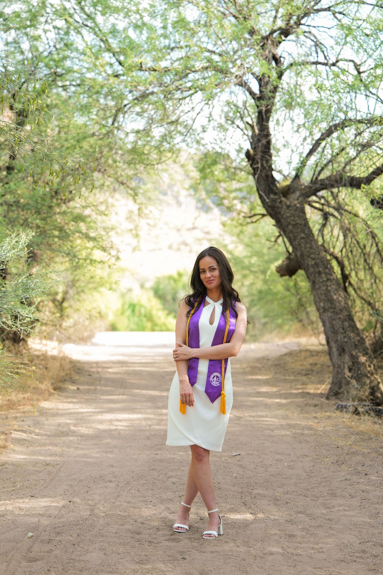 Young woman in white dress and purple graduation stole stands on a tree-lined dirt path for grad photos.