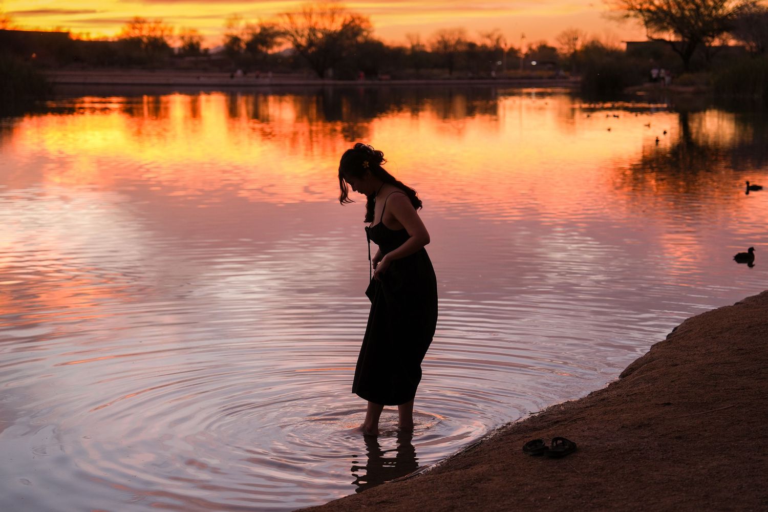 Silhouette stands at water's edge during golden sunset as orange sky reflects on calm lake surface.