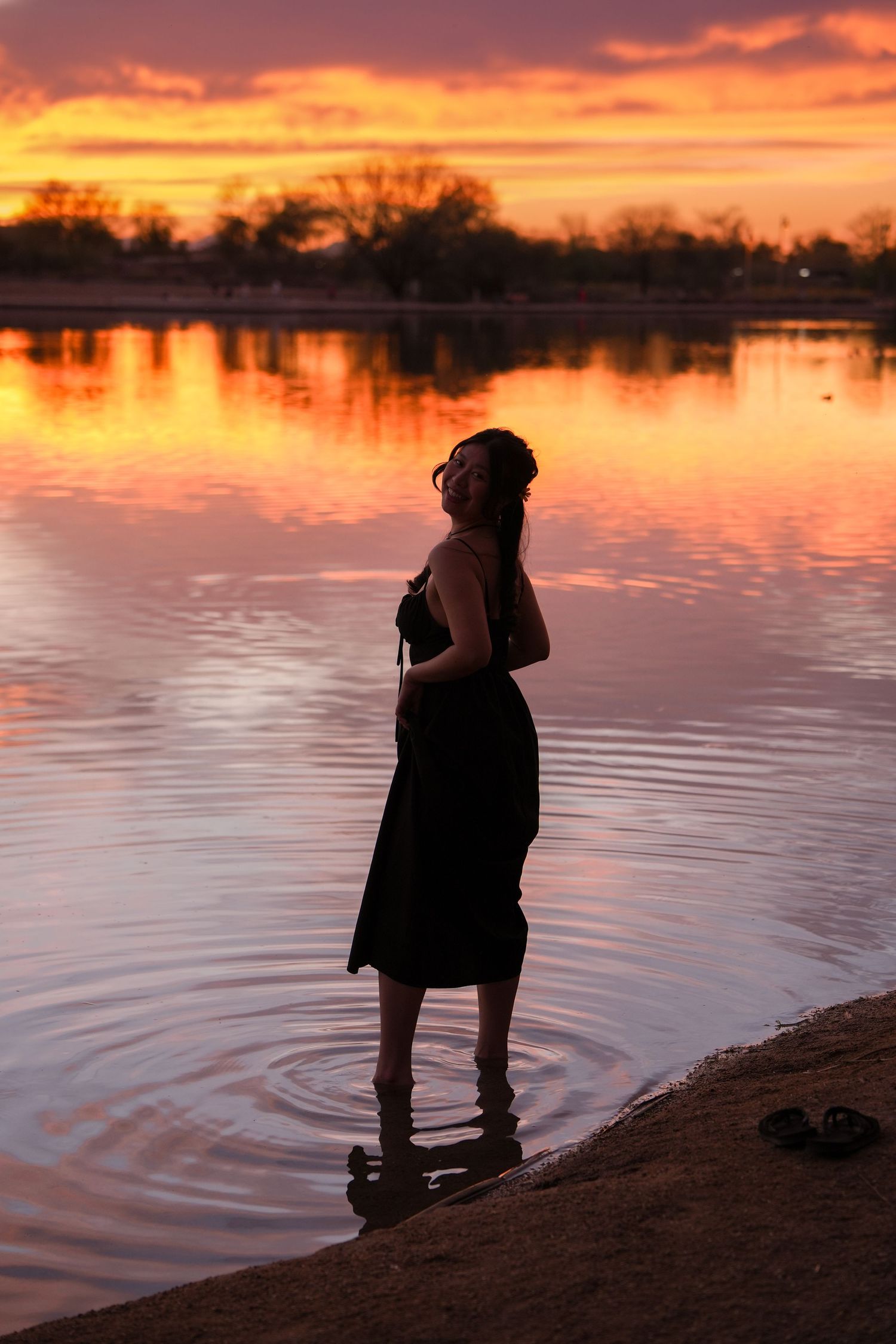 Silhouetted figure stands by tranquil lake at sunset with vibrant orange sky reflecting on calm water surface.