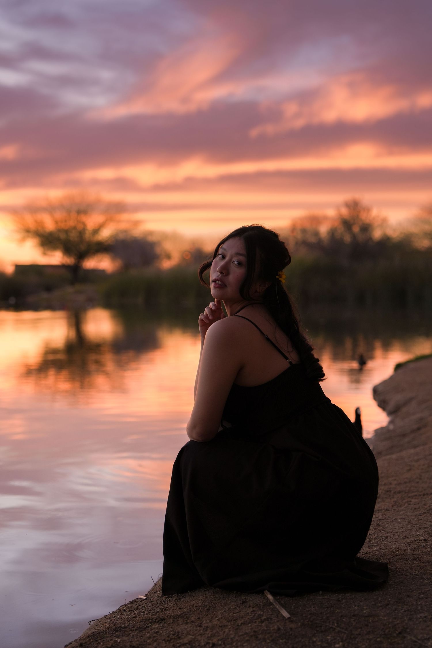A silhouette sits by a tranquil lake during a vibrant sunset with orange and purple clouds reflecting on the water.