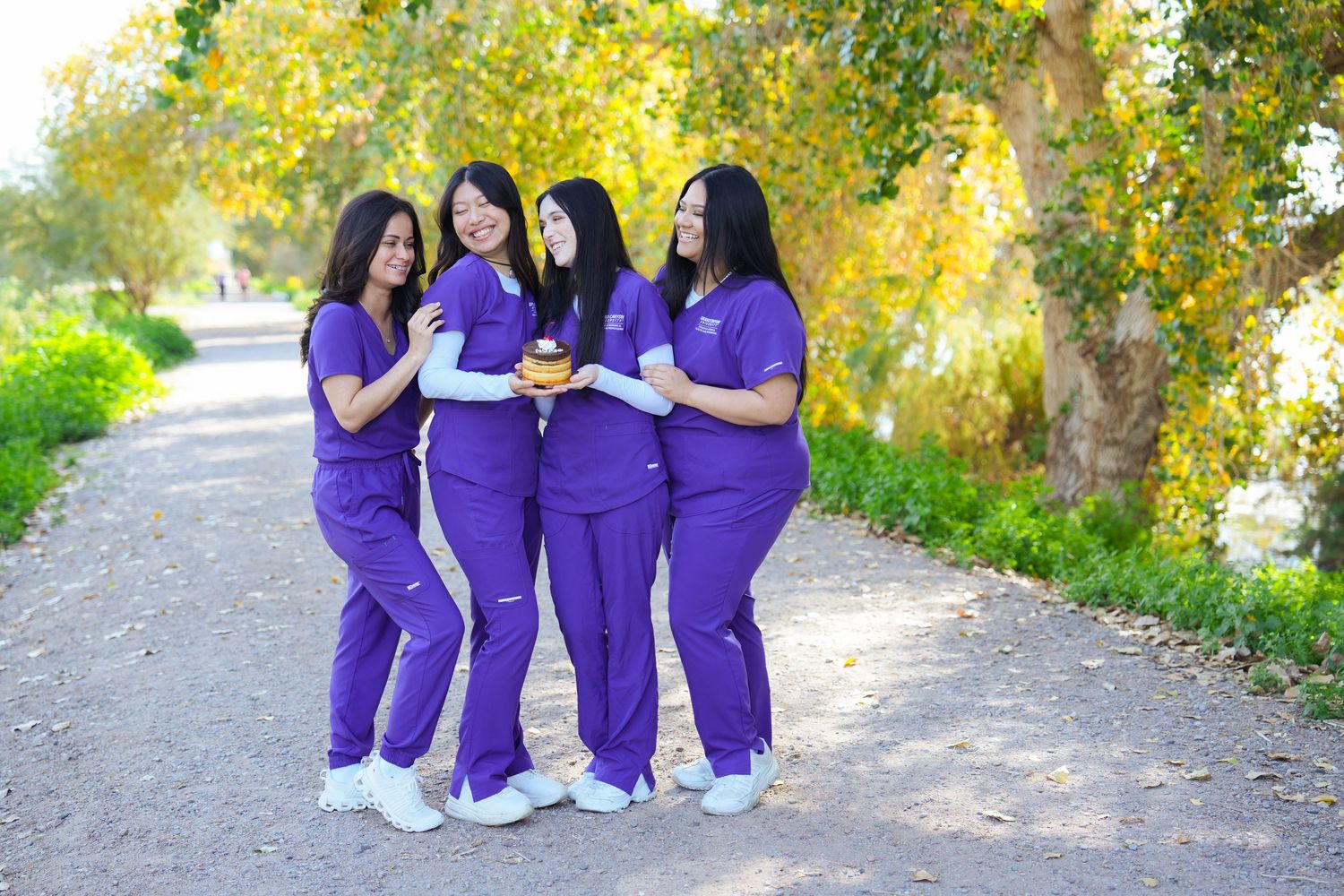 Healthcare workers in matching purple scrubs and white shoes stand together on an outdoor path surrounded by autumn foliage.