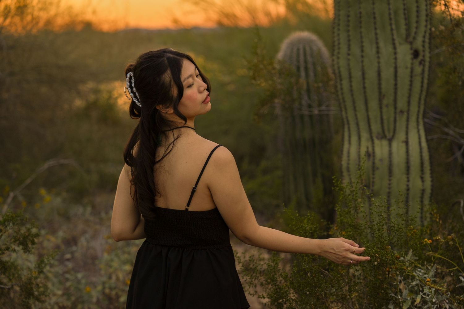 Young woman in black dress touching a saguaro cactus at golden hour sunset in Arizona desert.