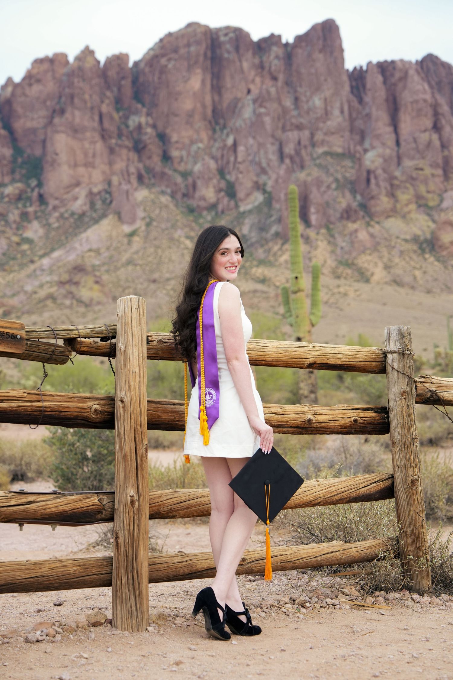 Young woman in white dress with purple graduation sash holds cap by rustic fence with Arizona desert mountains backdrop.