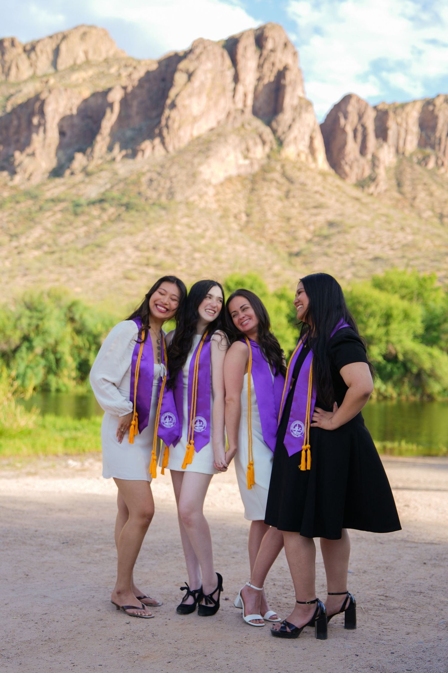 Four female graduates wearing purple stoles pose together outdoors with rocky mountains and a river in the background.