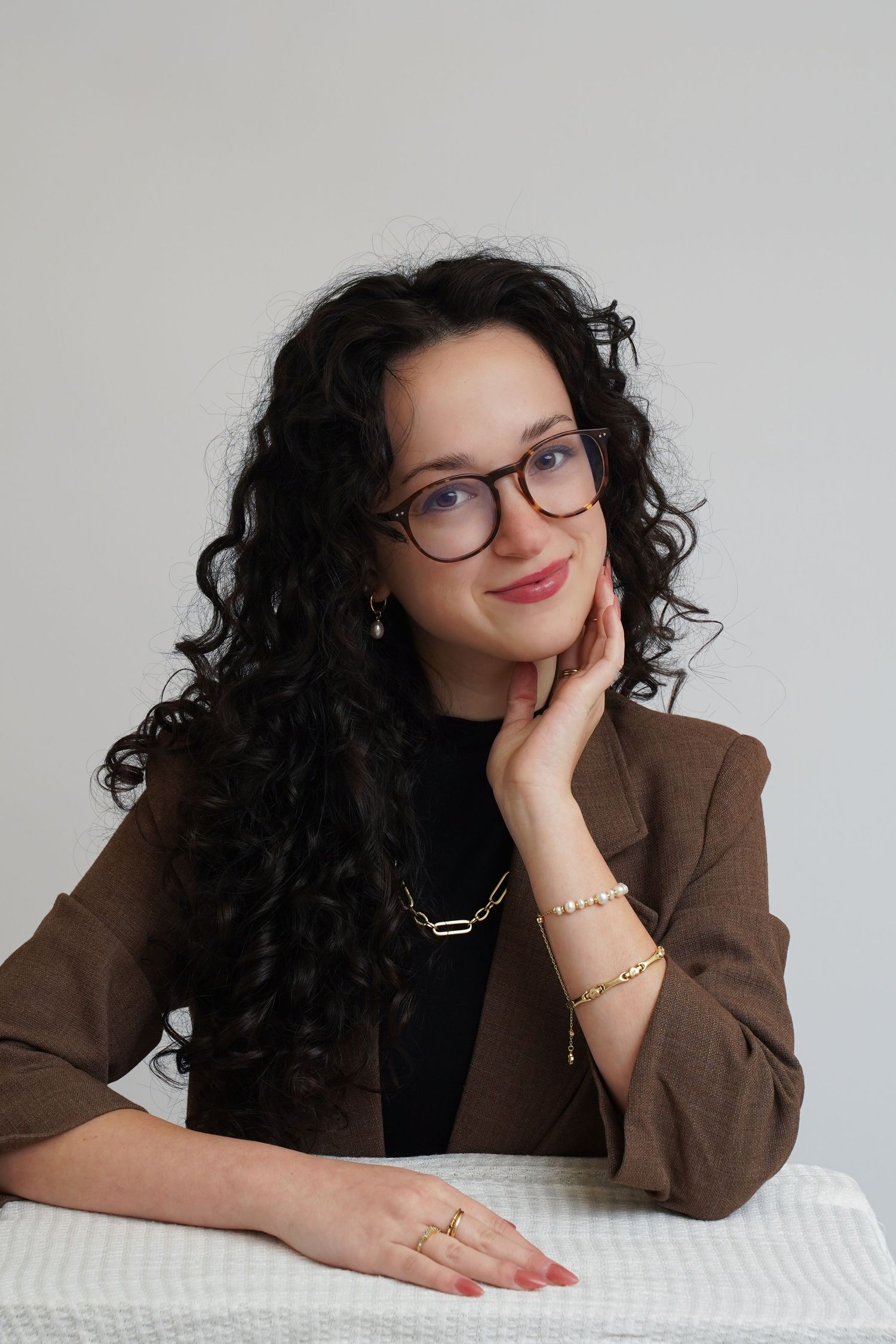 Professional headshots of someone with curly dark hair and glasses in a brown blazer against white background.