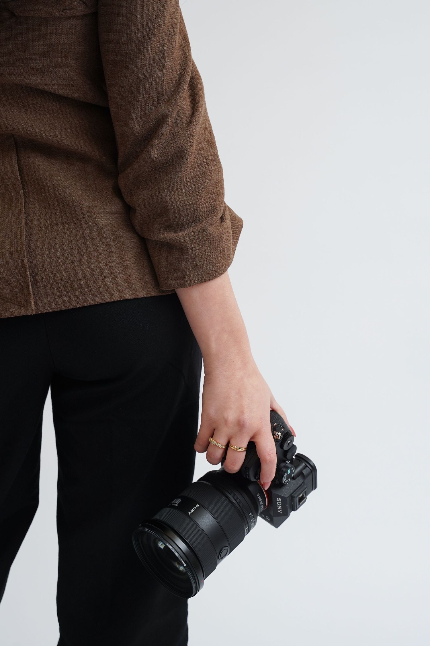 Person holding a professional DSLR camera with a large lens, wearing a brown blazer and black pants against a white background.