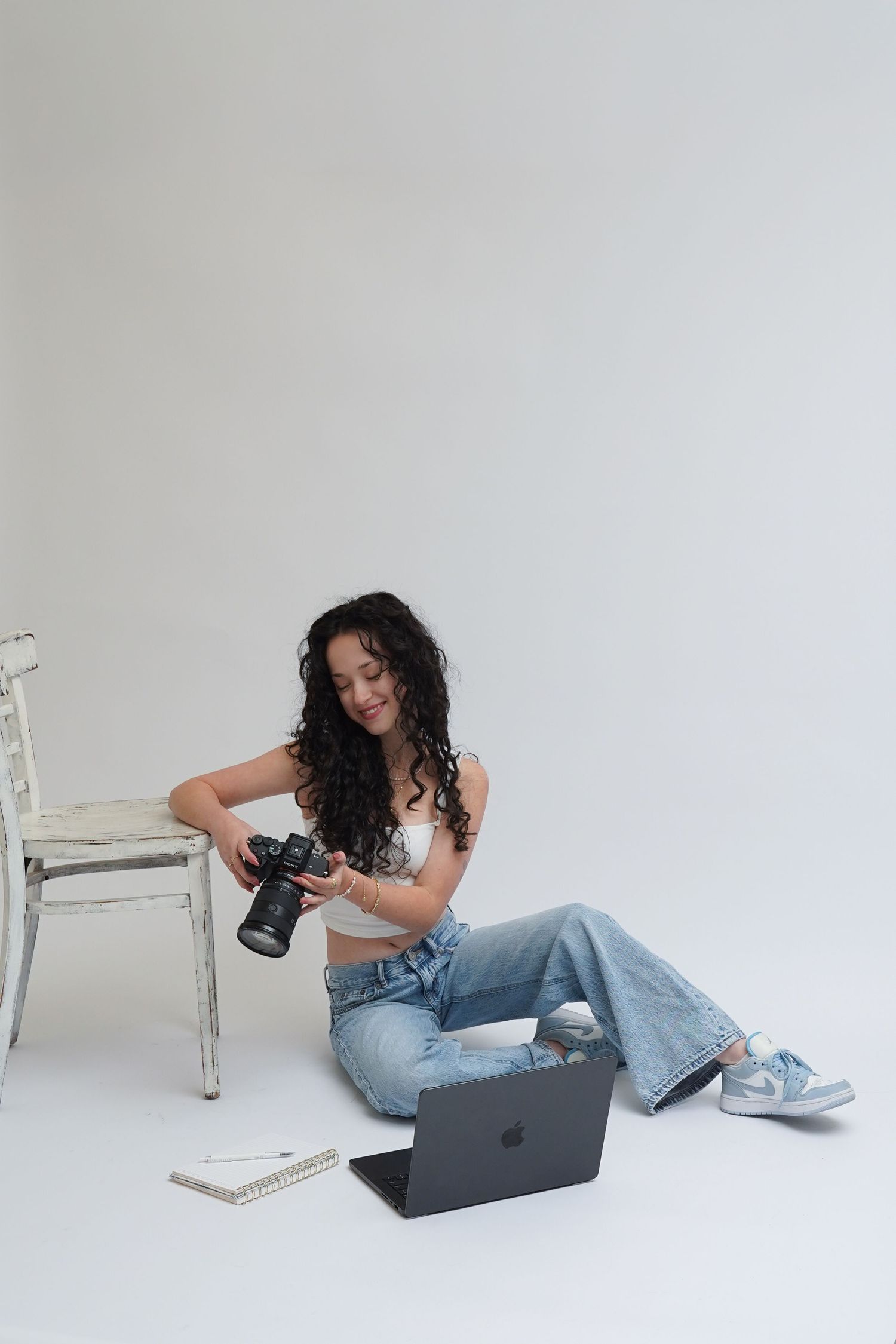Young woman with curly hair sits on floor holding DSLR camera beside laptop, wearing jeans and crop top in white studio.
