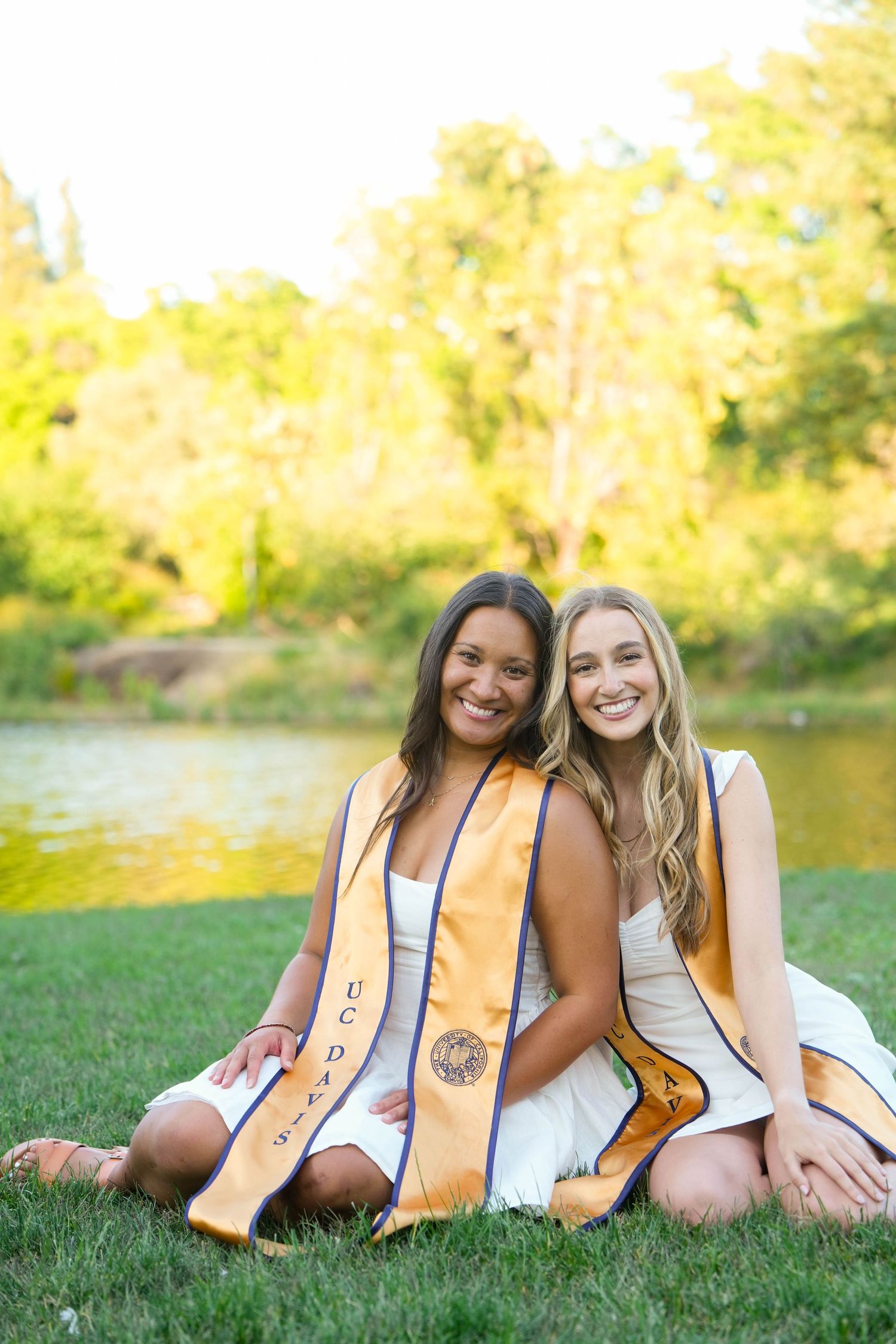 Two graduates in gold stoles sit together on grass by a pond during a sunny outdoor photo session.