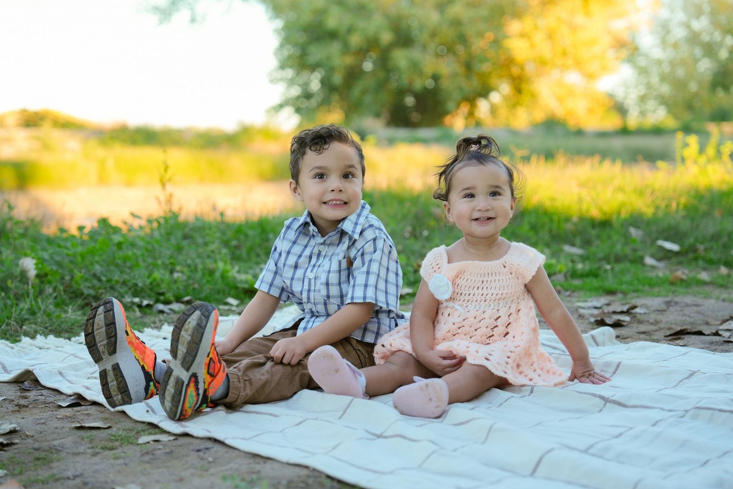 Children sitting on a white blanket outdoors surrounded by vibrant yellow wildflowers and green grass.