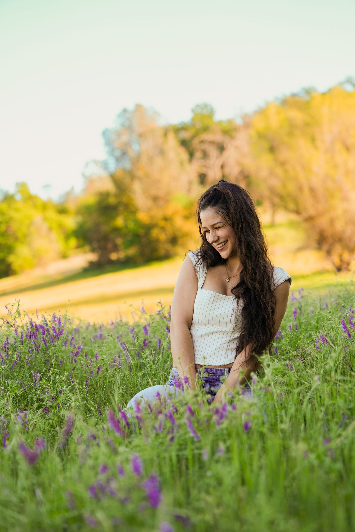Young woman with long dark hair smiles while sitting among purple wildflowers in a sunny green meadow.